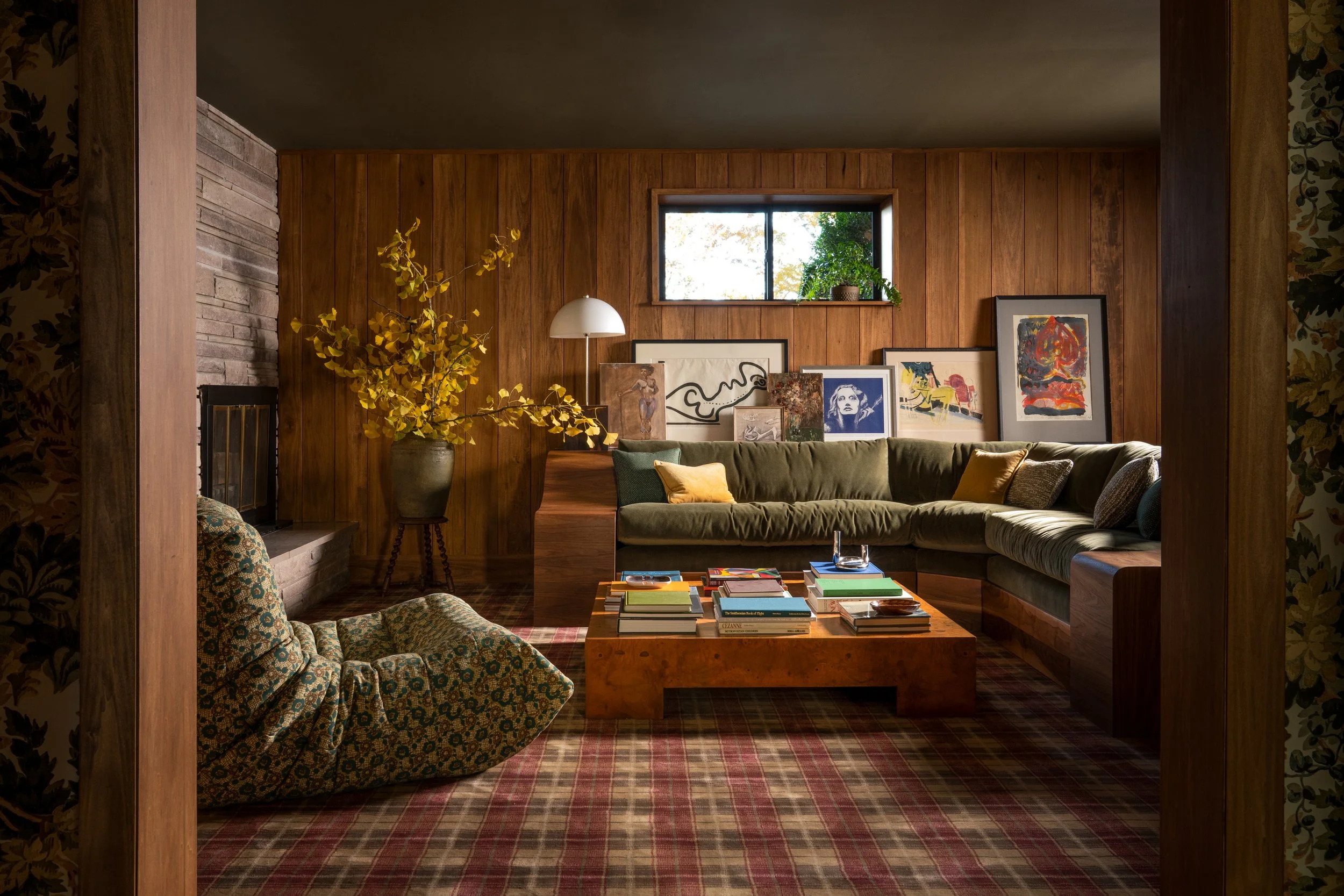 Cozy living room with wood-paneled walls, a large green sectional sofa with yellow and gray pillows, a vintage armchair with patterned fabric, a wooden coffee table with books, an artwork gallery on the wall, a window with greenery outside, a tall ye