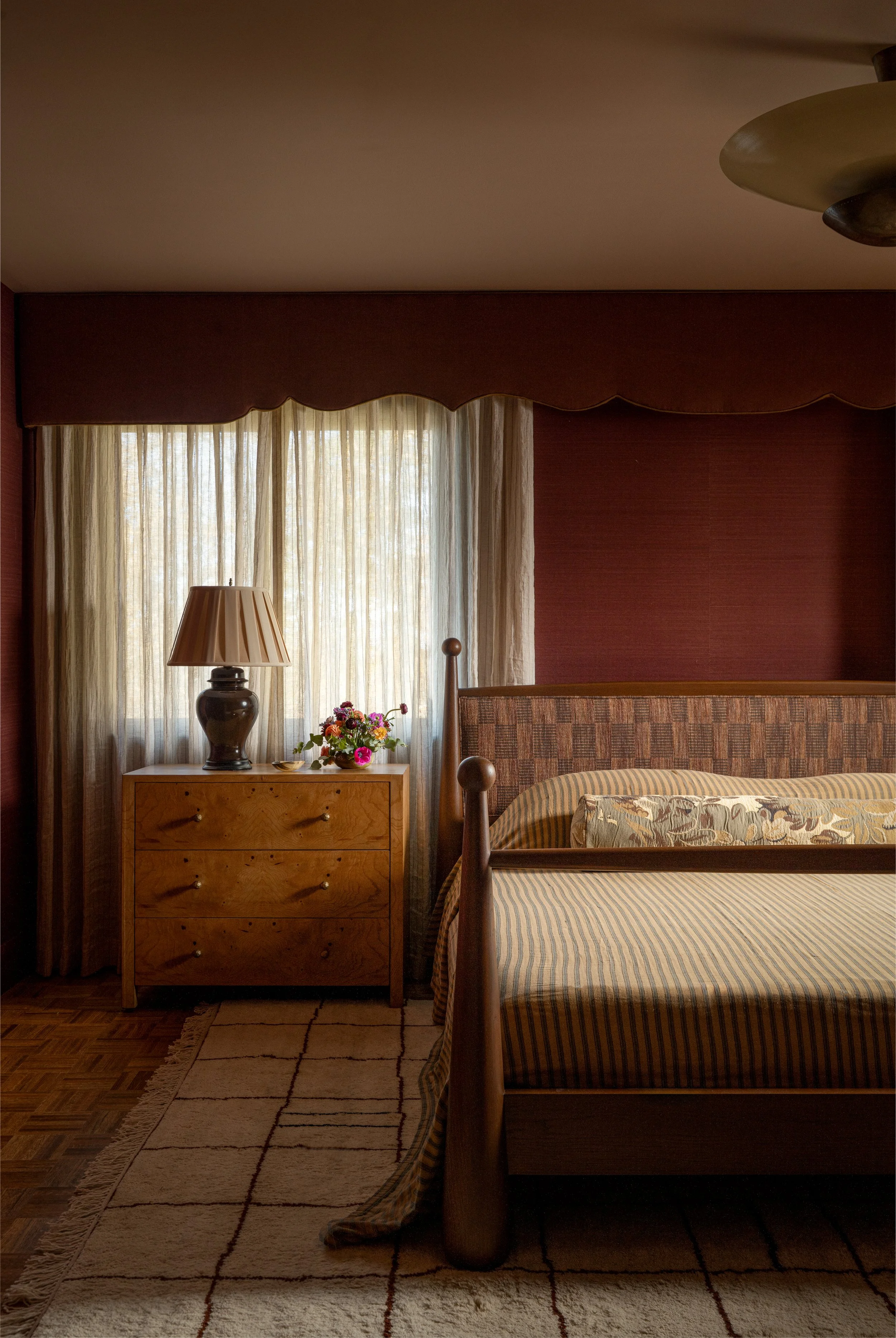 A cozy bedroom with a wooden bed, patterned quilt, floral pillow, wooden nightstand with lamp, and vase of colorful flowers, next to a window with curtains and a ceiling light fixture. Captured by Jeff Jones of Jeff Jones Photography, an interior pho