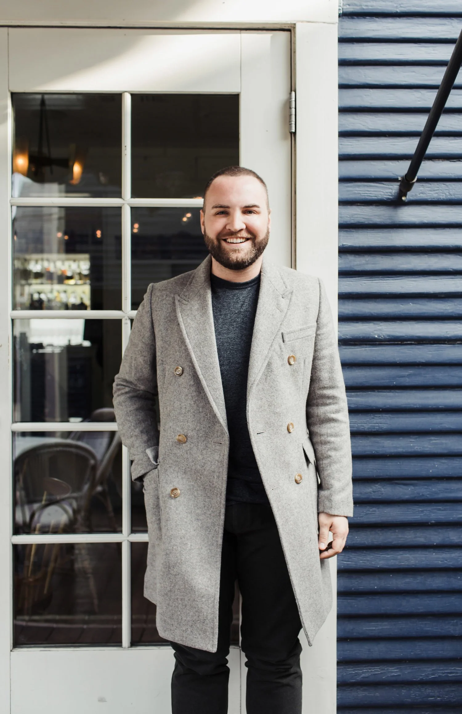 Taylor Murphy, A smiling man with a beard and short hair standing outside a building with a glass door and blue siding, wearing a grey coat and black pants.