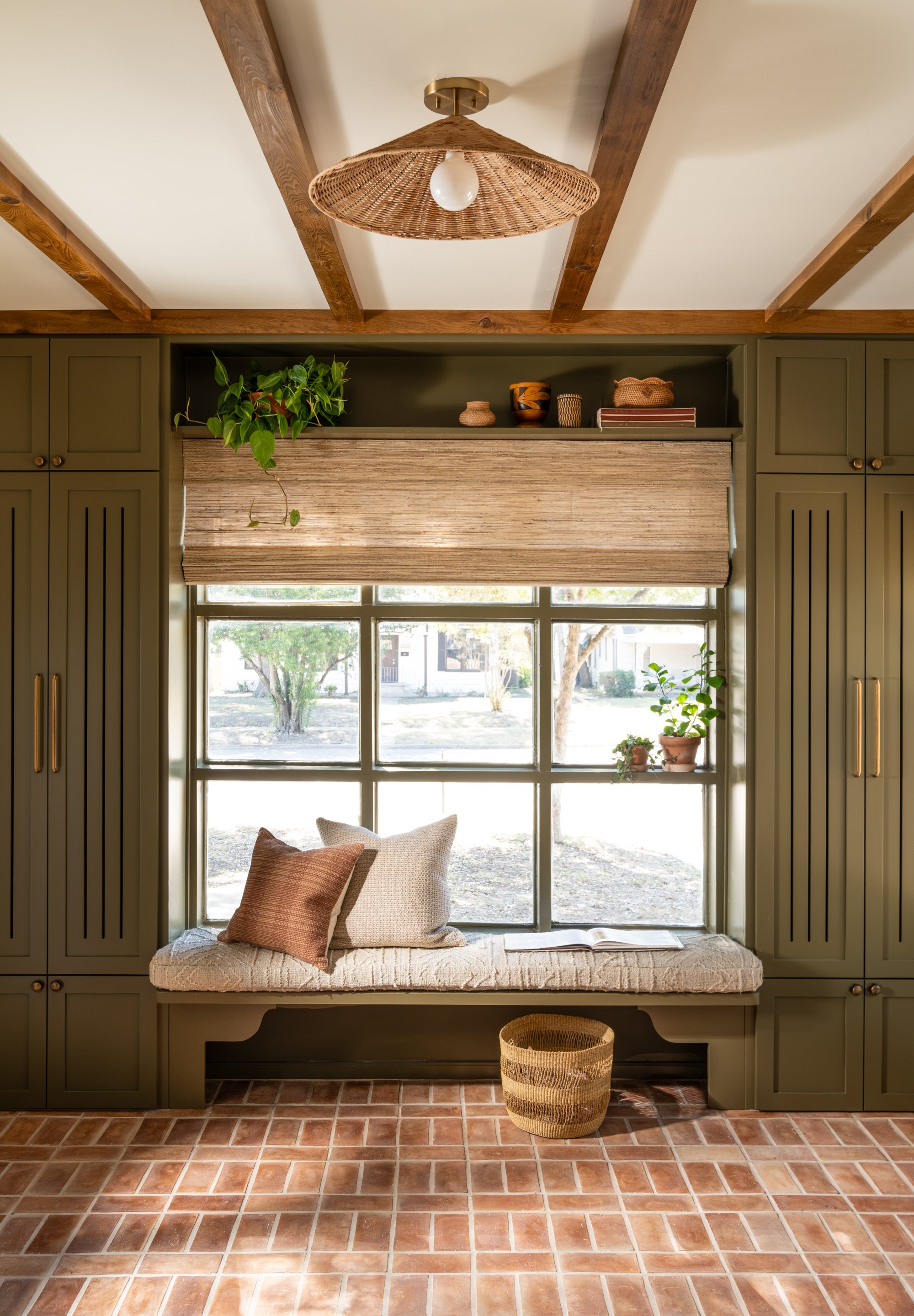 A cozy window nook with green cabinets on either side, a large window with a beige roller shade, a cushioned bench with pillows, a potted plant, a wicker basket, and a view of trees outside. The ceiling has wooden beams and a woven pendant light. Cap