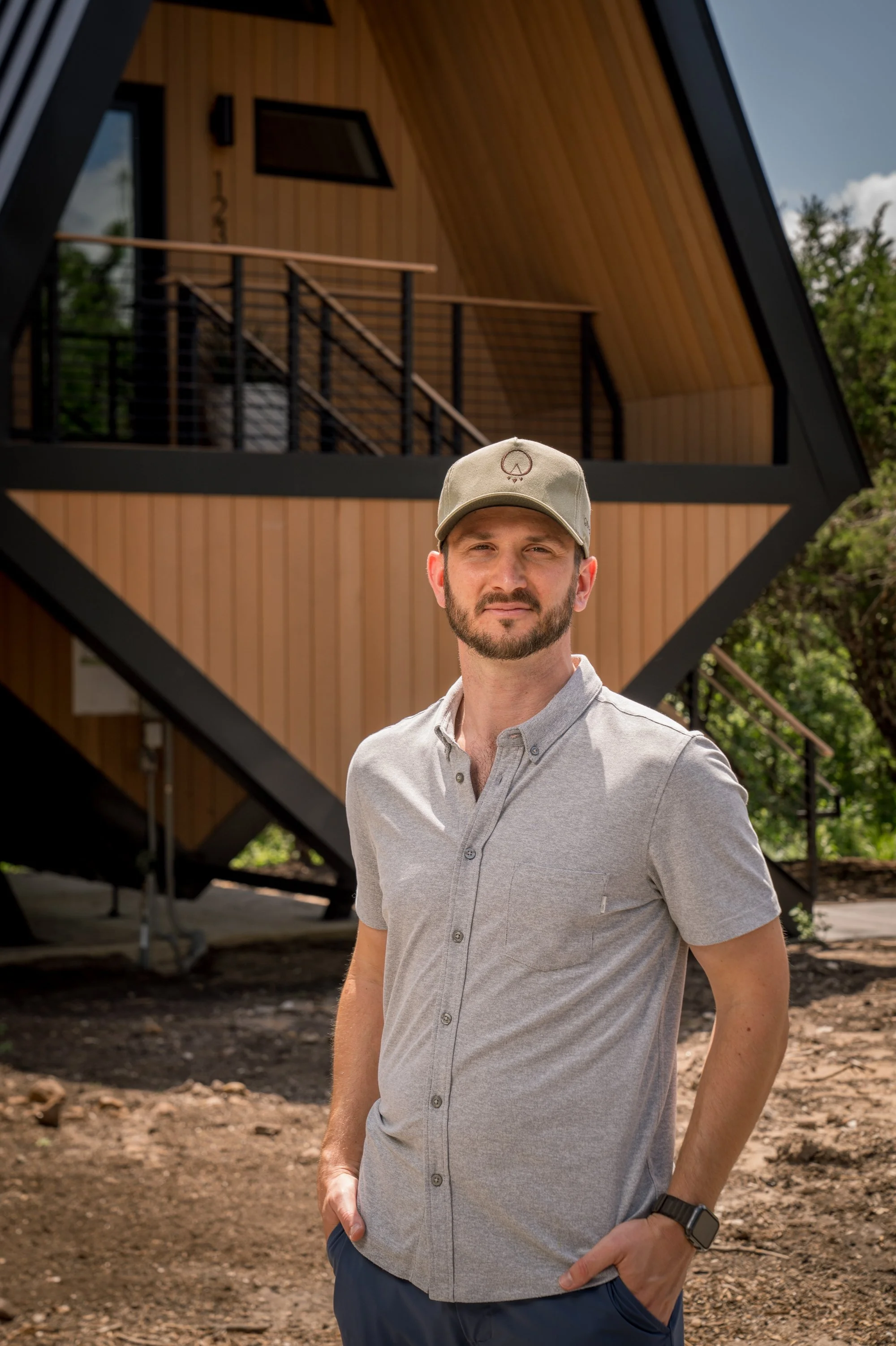 A man with a beard wearing a gray button-up shirt, a beige cap with a peace symbol, and a black wristwatch standing outdoors in front of a modern A-frame wooden house with large windows and a staircase leading to a balcony.