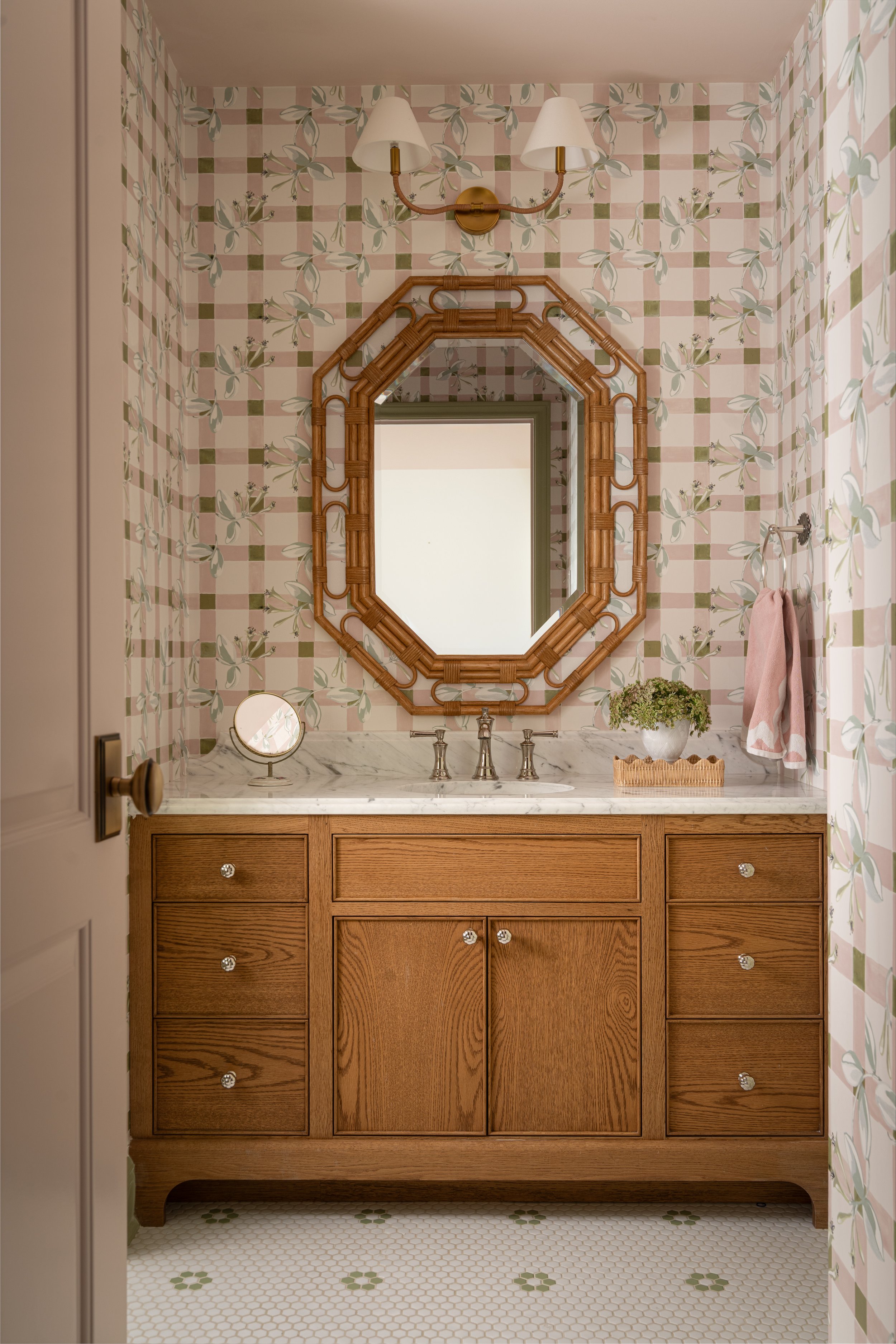 A bathroom with floral wallpaper, a wooden vanity with a marble countertop, a decorative wooden mirror, a wall-mounted light fixture, a small round mirror, a potted plant, and a pink towel. Captured by Jeff Jones of Jeff Jones Photography, an interio