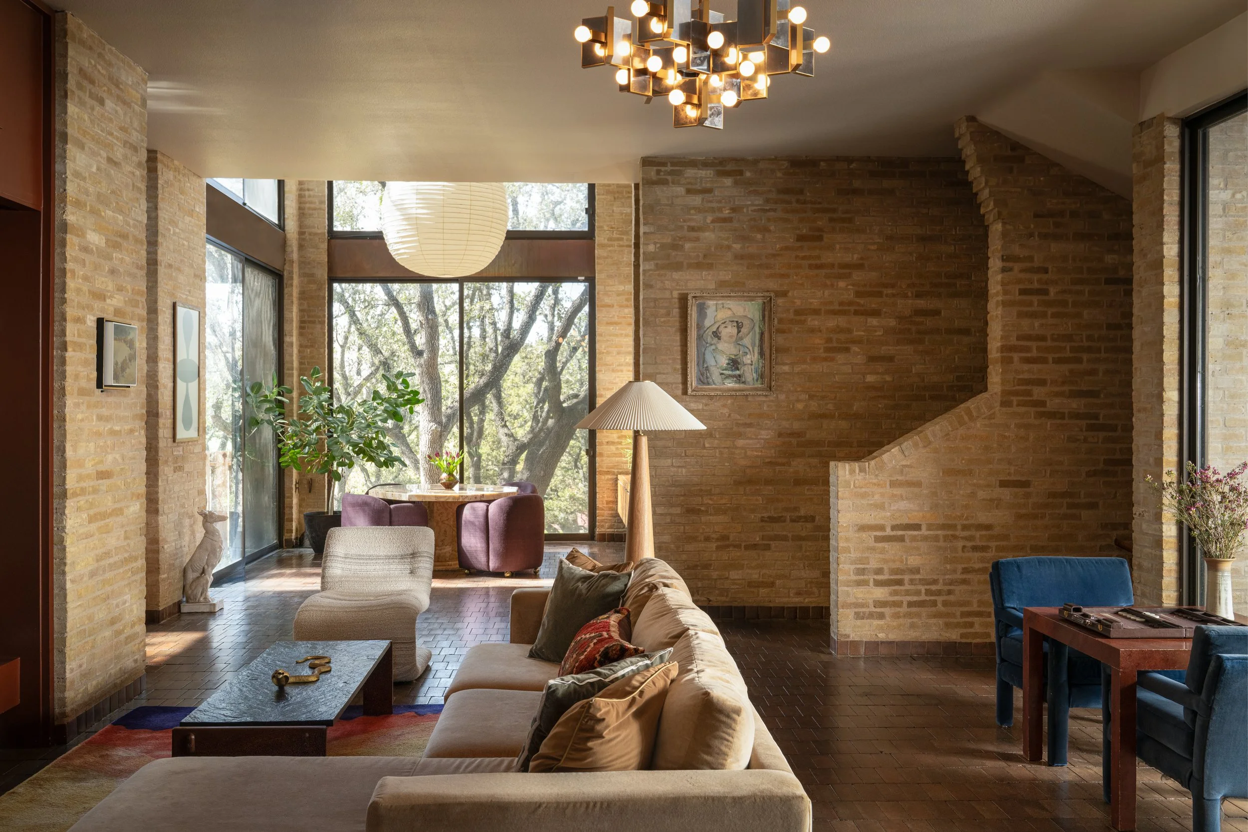 Living room with exposed brick walls, large windows, a beige sofa with pillows, a white armchair, a wooden coffee table, a tall lamp, a dining table with purple chairs, and artwork on the walls. There's a large plant and a view of trees outside. Capt