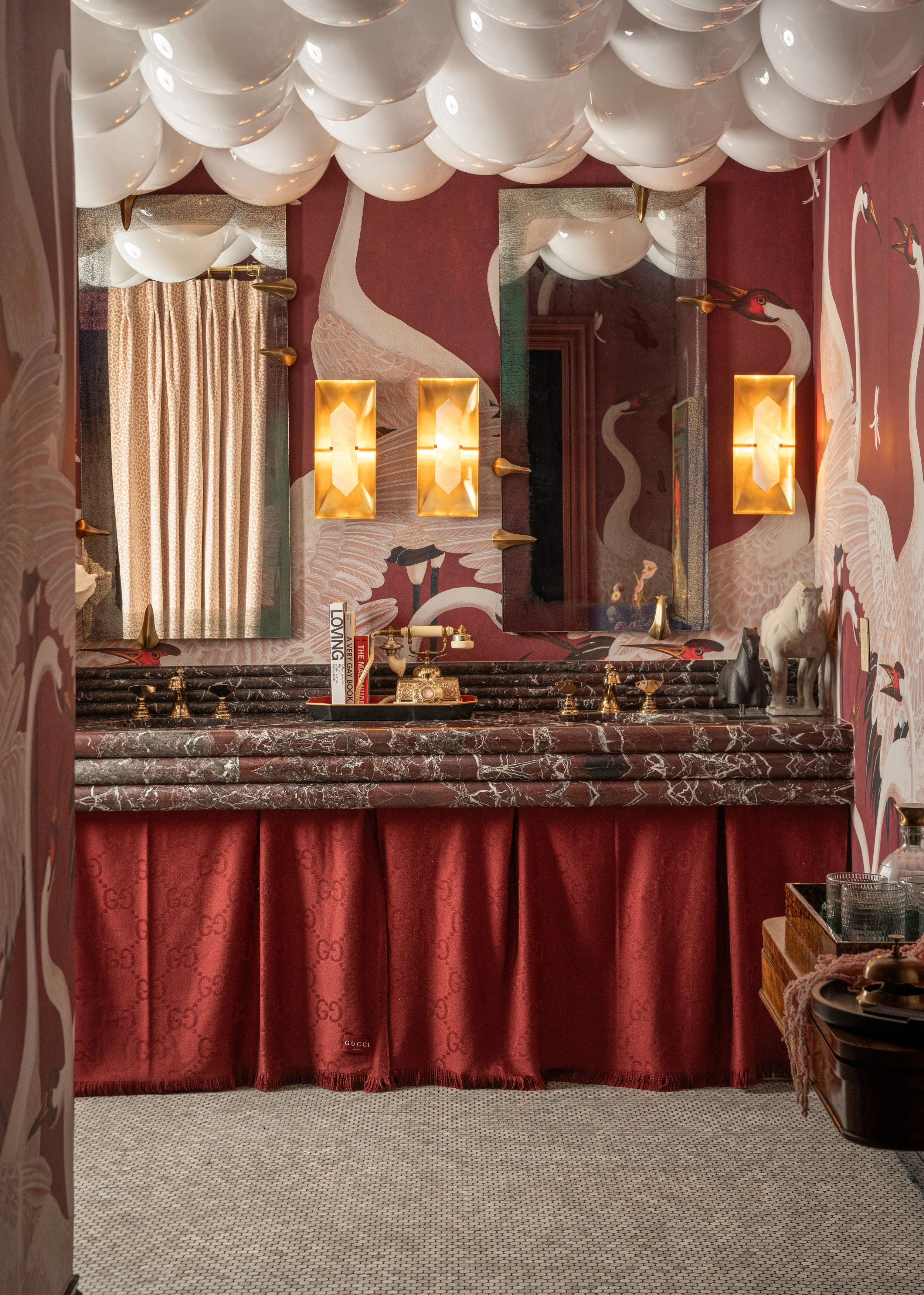 A bathroom with red wallpaper featuring white and black heron illustrations, two rectangular gold wall sconces, two large mirrors, a marbled countertop, decorative items including books, a vintage telephone, and animal figurines, with a red curtain u