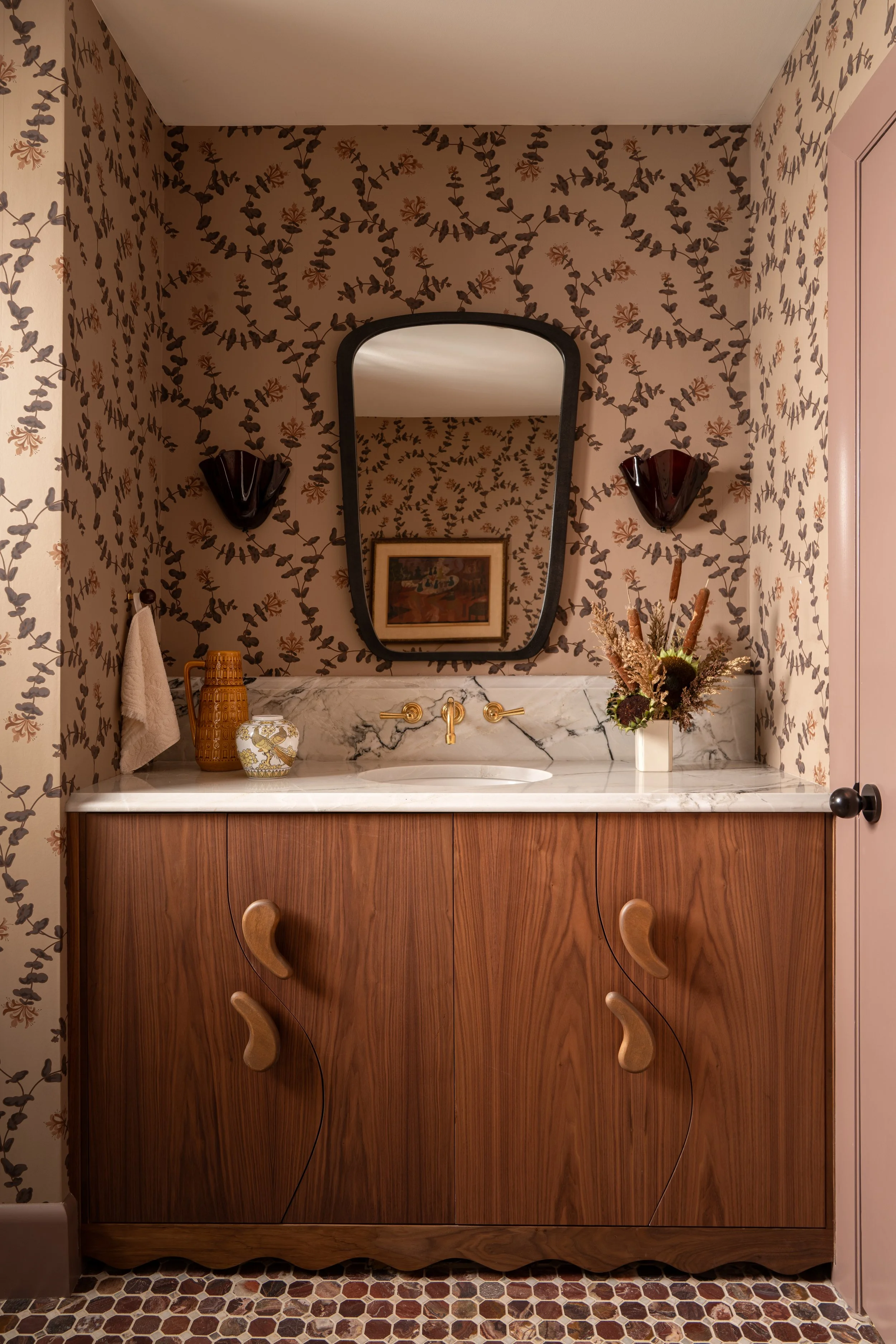 A vintage bathroom vanity with a marble countertop, a decorative vase, a wall-mounted mirror, floral wallpaper, a wall lamp, and a potted flower arrangement. Captured by Jeff Jones of Jeff Jones Photography, an interior photographer based in San Anto
