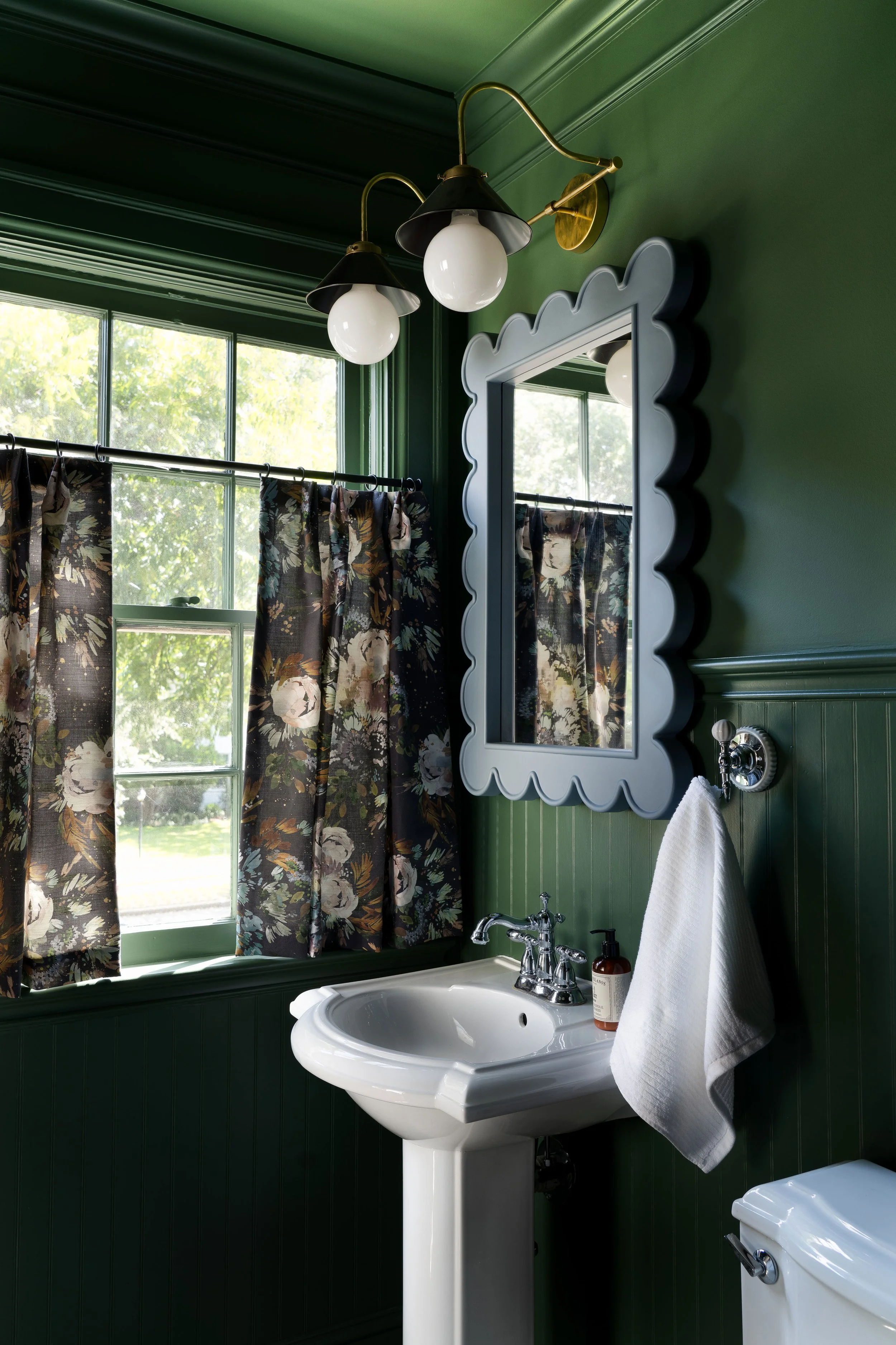 Interior design photography by Jeff Jones capturing a Dream Dutch Colonial bathroom by Whitney Walker on Farrington Lane. Features cafe curtains, scalloped mirror, brass lights, and dark green walls in Waco, Texas.