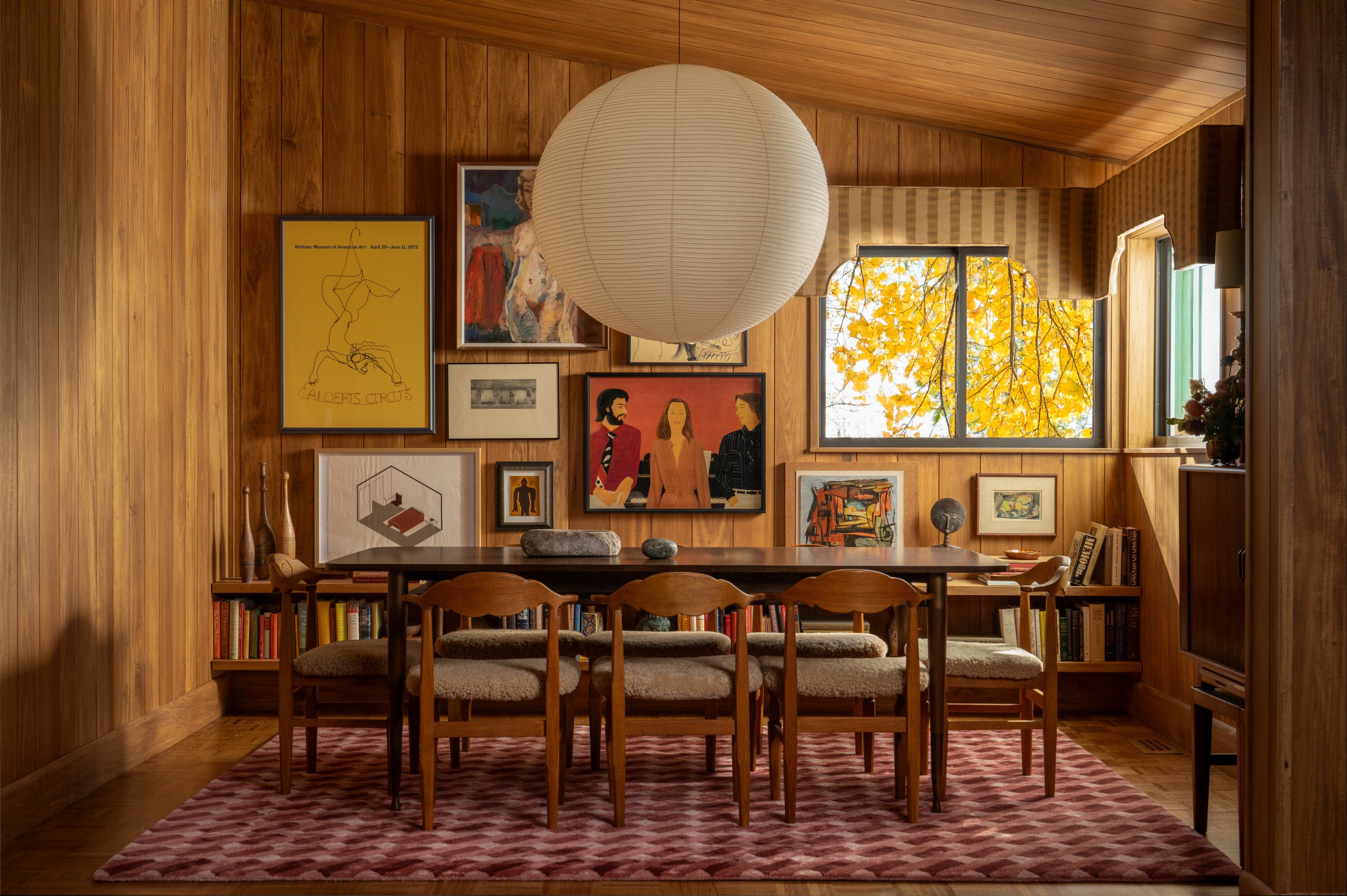 A cozy dining room with wooden walls and ceiling, a large paper lantern hanging from the ceiling, a dark wood dining table with six upholstered chairs, a patterned pink area rug, and a gallery wall of framed artwork and photographs. The room has a wi