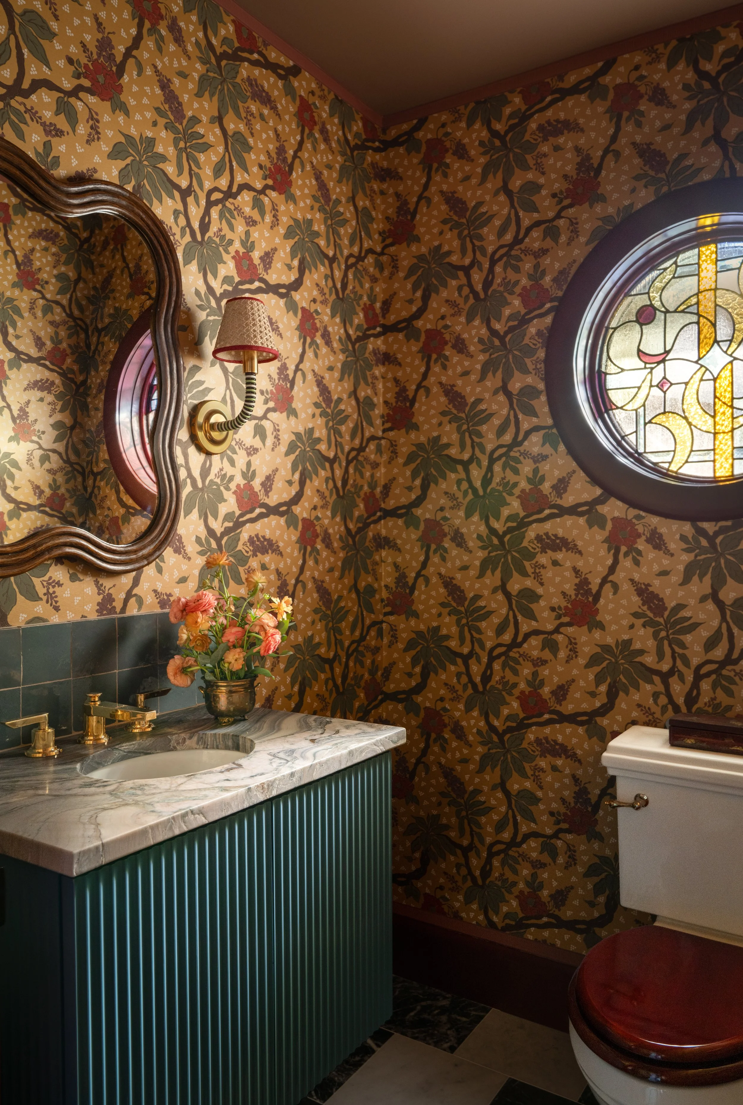 A vintage-style bathroom with floral wallpaper, a marble countertop sink, a green vanity with vertical lines, a brass faucet, a small flower arrangement, a decorative wooden framed mirror, a wall-mounted lamp with a fabric shade, an oval stained glas