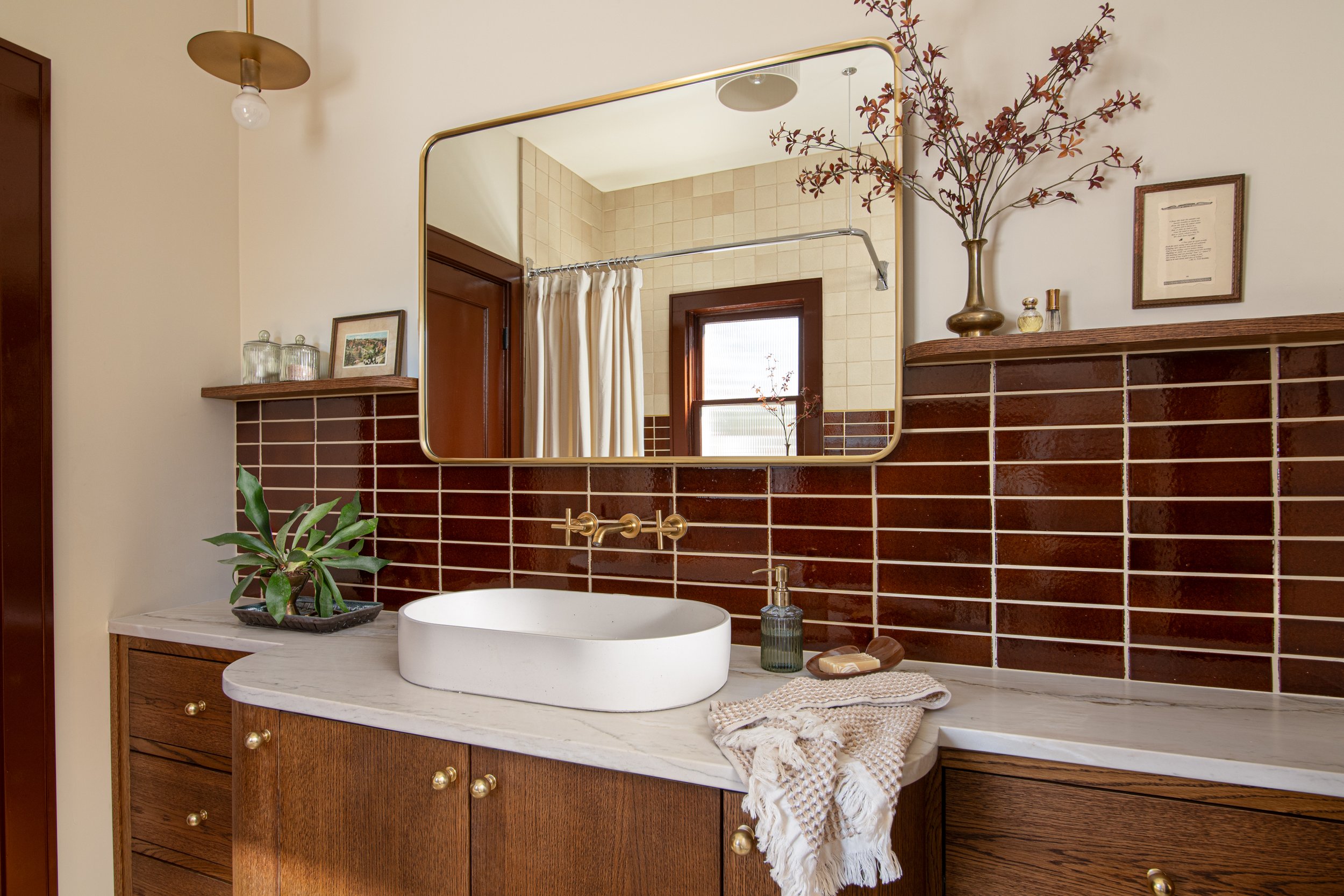 Bathroom vanity with a marble countertop, a white vessel sink, a soap dispenser, a small plant, and decorative items. A large mirror and a wooden shelf with glass jars and a framed photo. Brown tile backsplash and a window with curtains. Captured by 