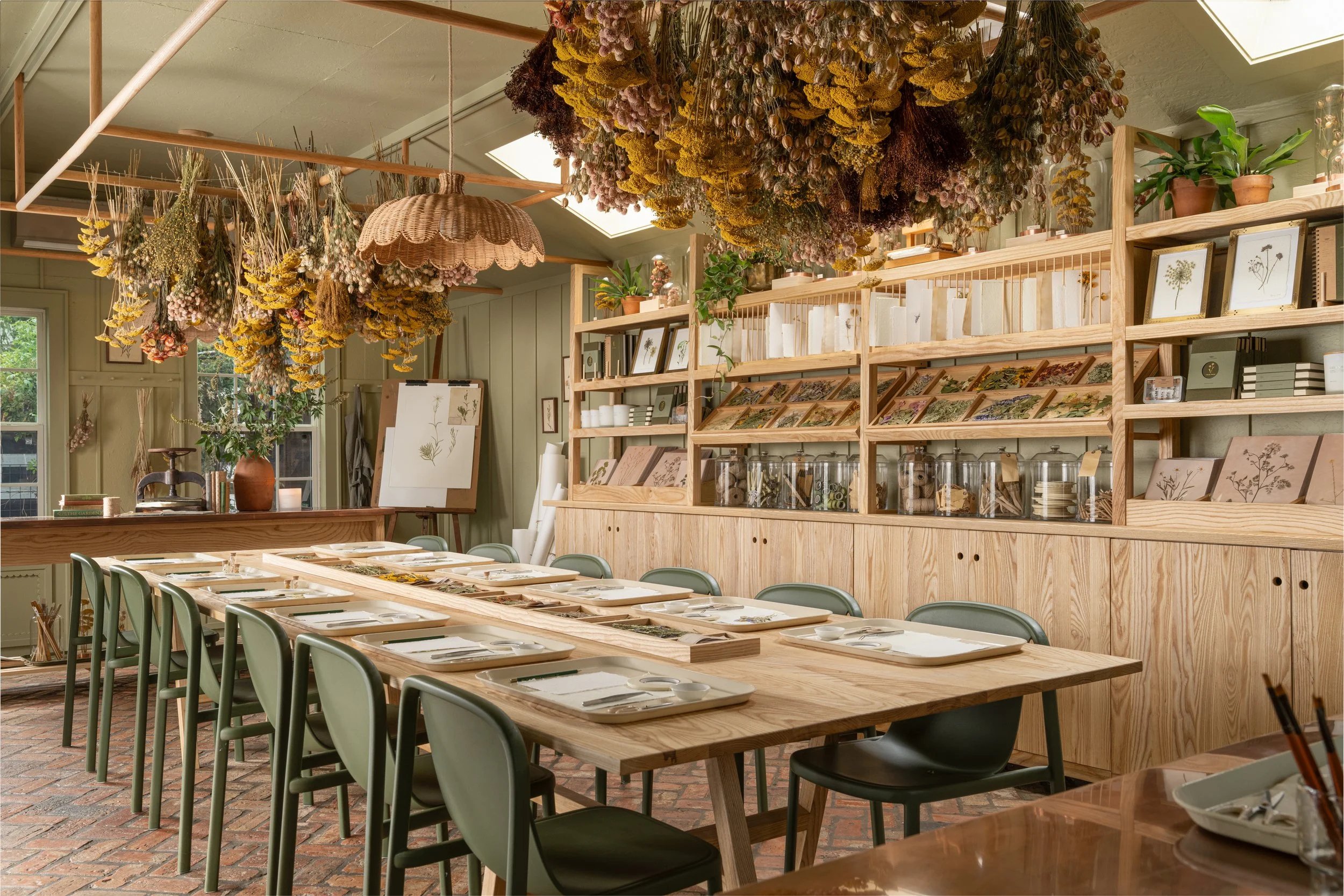 A cozy dining room with a long wooden table set with white plates, napkins, and cutlery, surrounded by green chairs. Overhead, there are numerous dried flower arrangements hanging from the ceiling. The background features wooden shelving filled with 