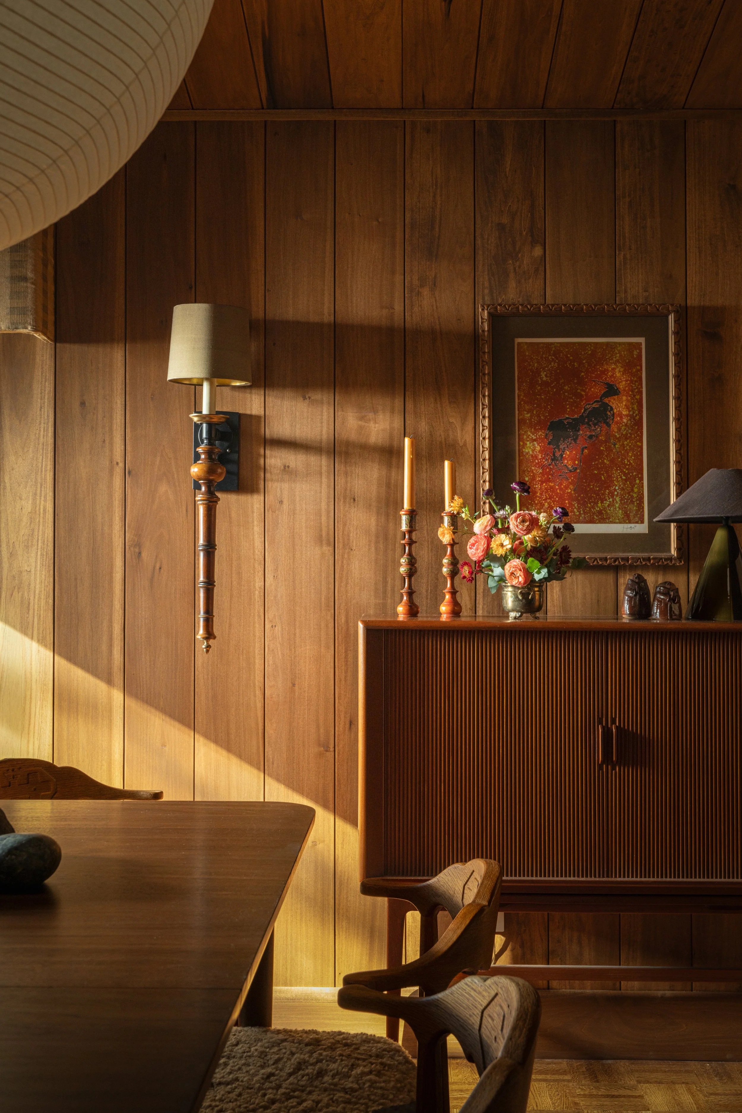 A wood-paneled interior room with a mounted lamp, a framed colorful artwork, a floral arrangement, three candlesticks, and small decorative items on a wooden sideboard. Captured by Jeff Jones of Jeff Jones Photography, an interior photographer based 