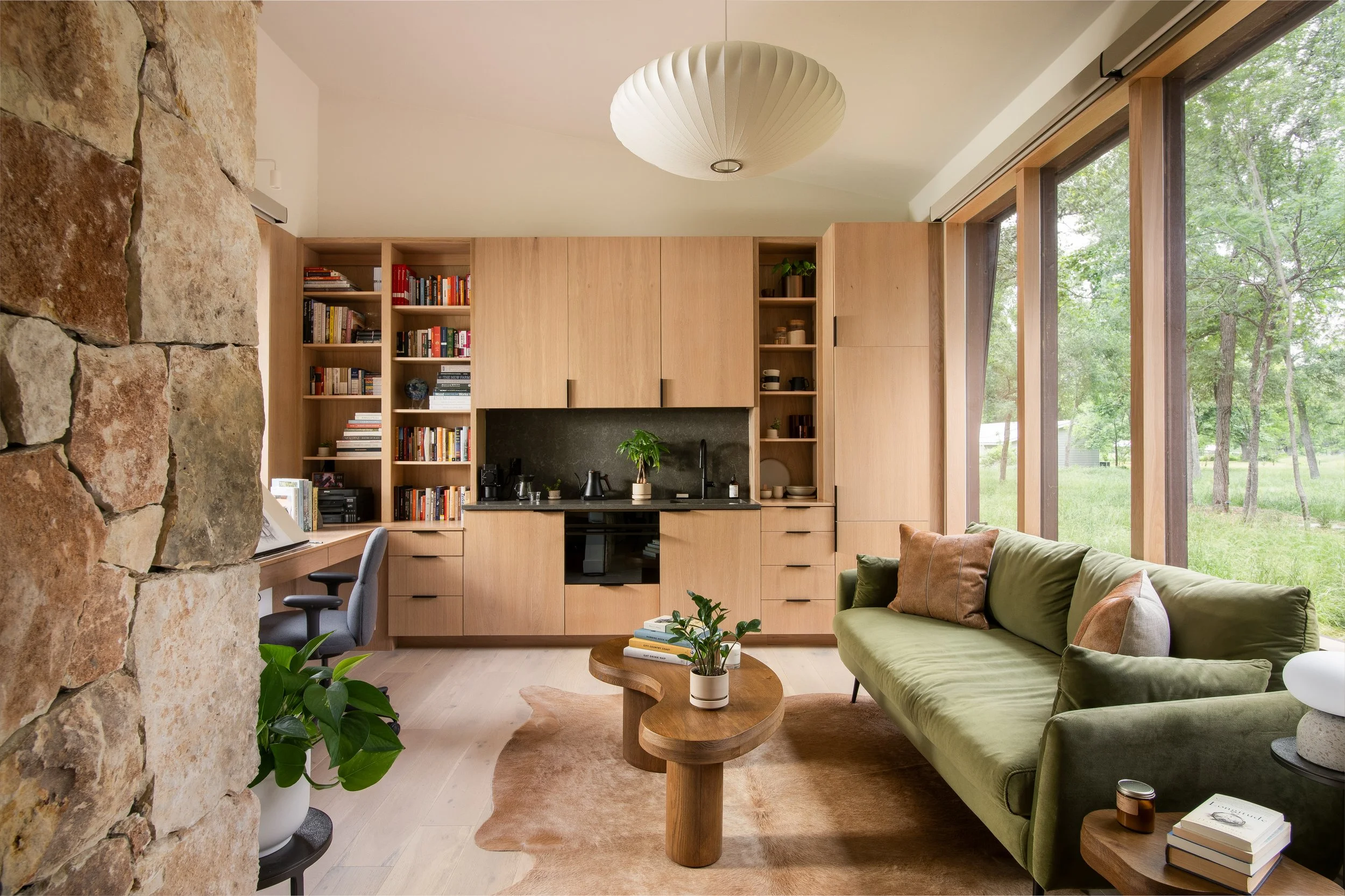 Interior design photography by Texas-based photographer Jeff Jones, featuring a cozy modern living space with wood cabinetry, green sofa, stone wall, and a large windows overlooking nature.