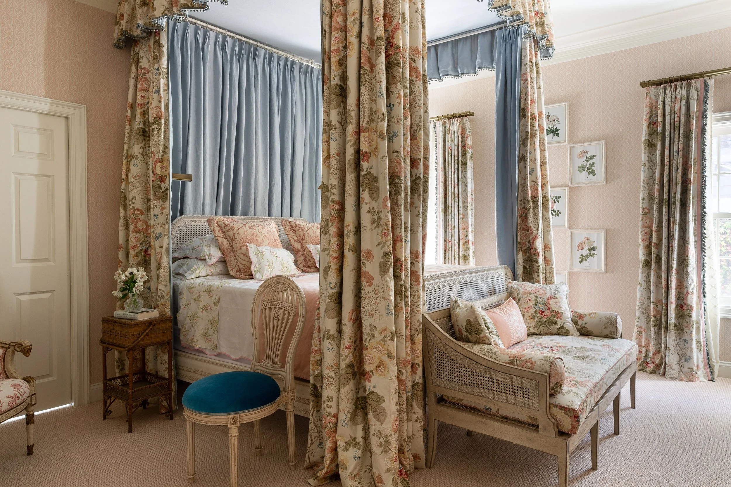 A soft, elegant bedroom featuring floral drapes framing a canopy bed, with light blue and neutral tones throughout, complemented by vintage furniture pieces and delicate details by Mark D. Sikes Interior Photography