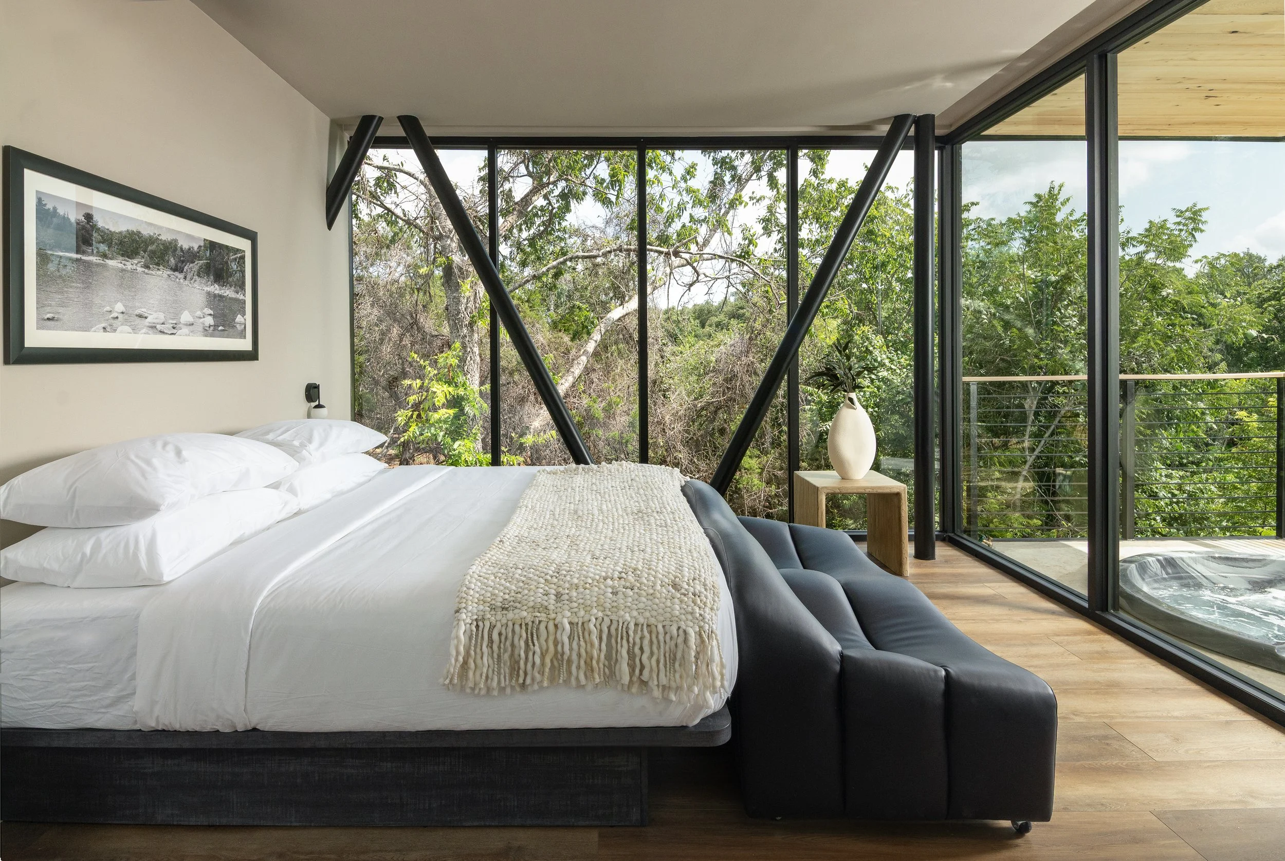 Modern bedroom with large glass windows, overlooking trees, with a bed, a black frame, white bedding, and a black couch at the foot of the bed. Captured by Jeff Jones of Jeff Jones Photography, an interior photographer based in San Antonio, Texas