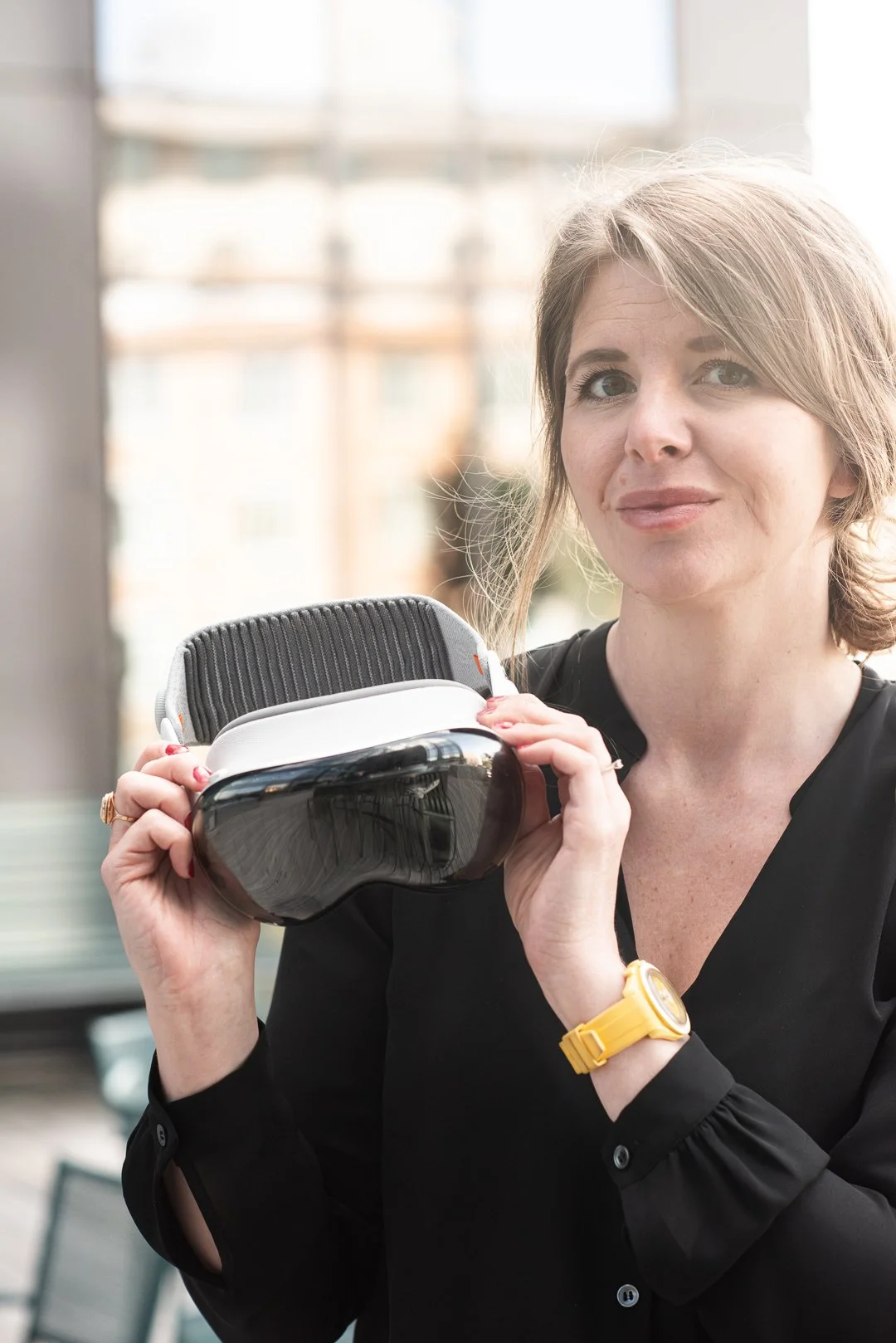 Ung kvinna som bär en svart blus och gul armbandsklocka, håller en VR-headset i handen och ser mot kameran på en ljus, urban bakgrund.