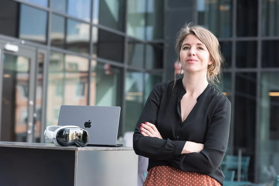 Kvinna står utomhus framför en modern glasbyggnad, med korsade armar, svart blus och rödbrunt kjol med vita prickar, med en Apple MacBook-dator och en nytillverkad VR-glasögon på ett bord.