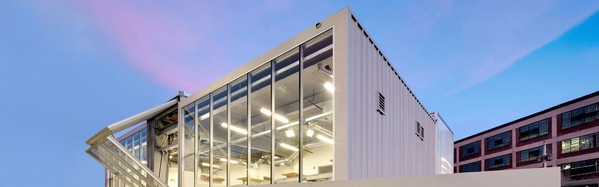 Modern building with large glass windows and illuminated interior, set against a sky with pink and blue hues