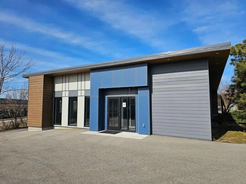 Modern single-story commercial building with glass doors, wood paneling, blue and gray siding, and a sloped roof on a paved lot under a blue sky.