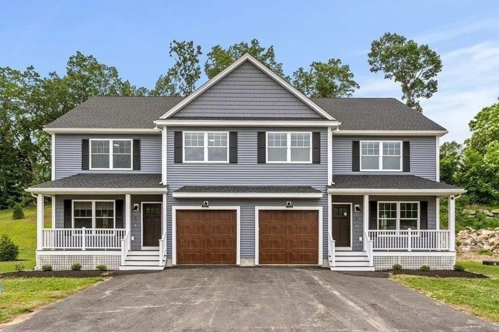 A modern duplex house with two attached units, each with its own garage, porch, and front door. The house has gray siding, white trim, and black shutters, with a landscaped yard and a paved driveway.