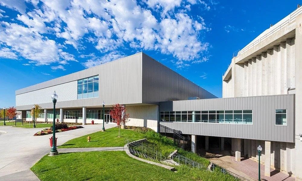 Modern building with steel siding, glass windows, a landscaped area with trees, flowers, and lamp posts, under a blue sky with scattered clouds.