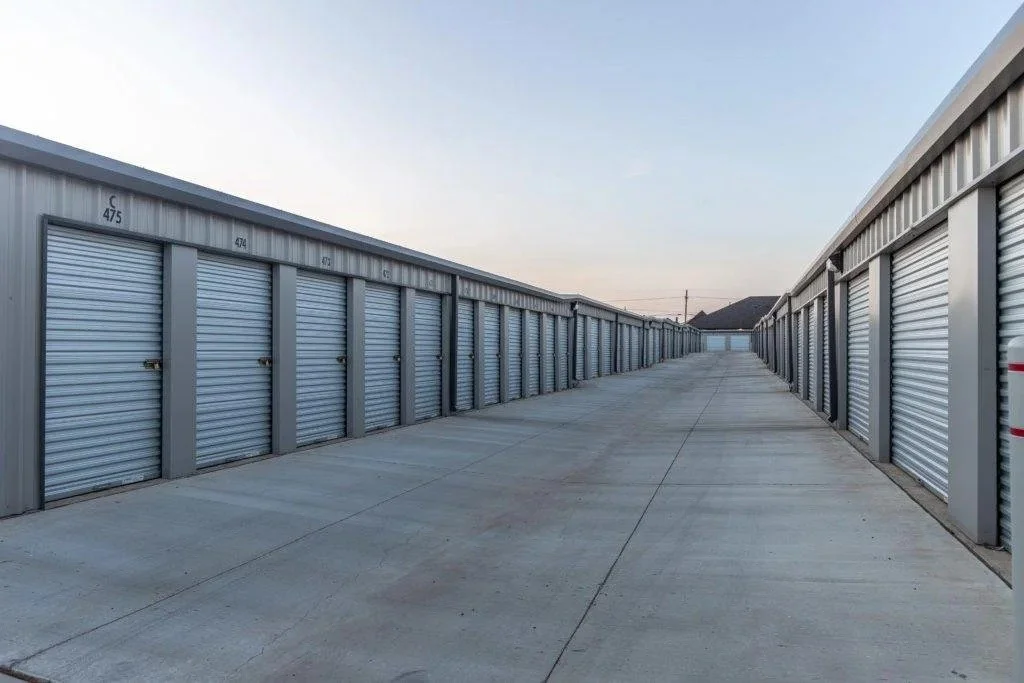 Row of storage units with closed metal roll-up doors along a concrete driveway, under a clear sky at dusk.