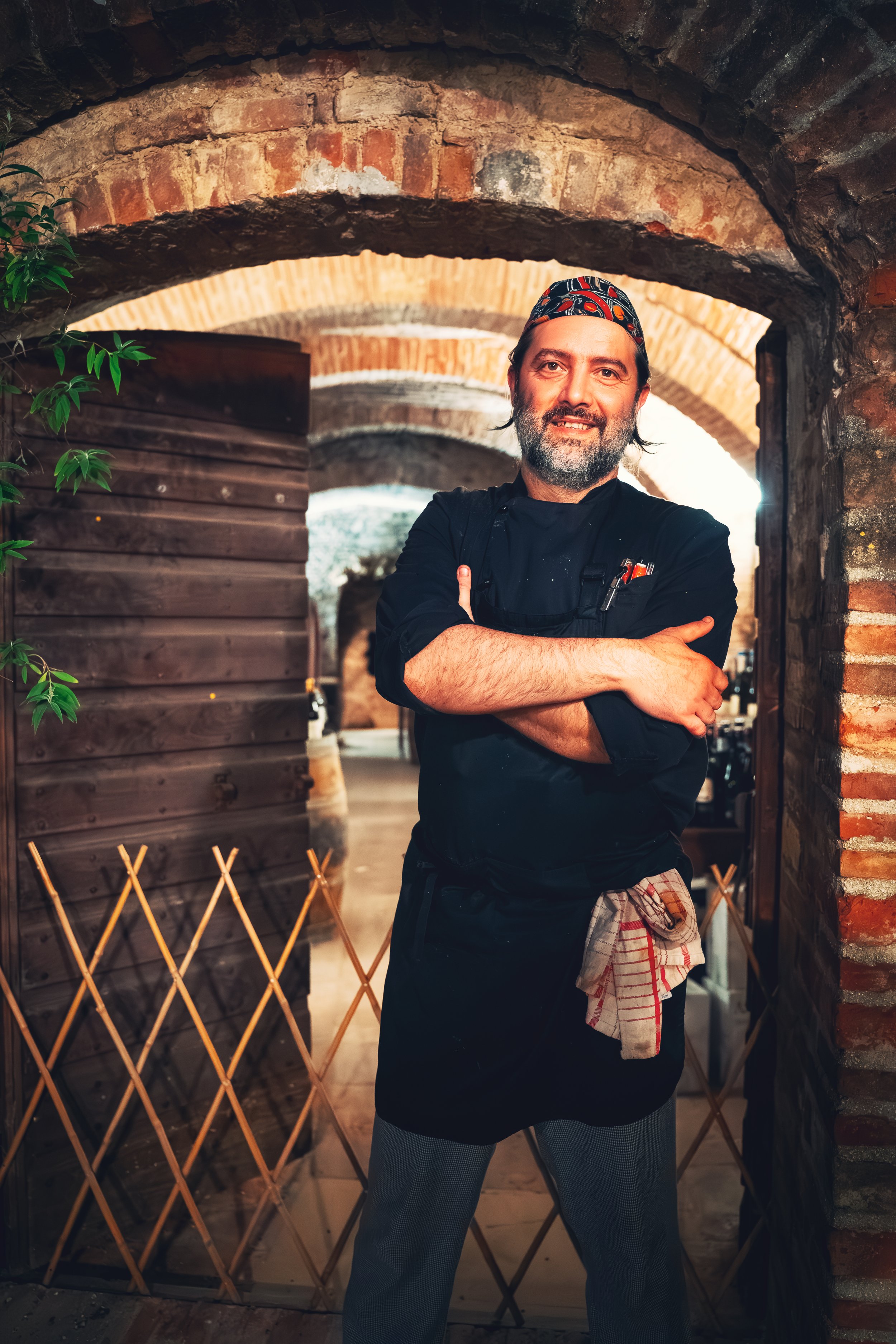 Chef standing in a brick archway, wearing a black apron and headscarf, smiling with arms crossed.