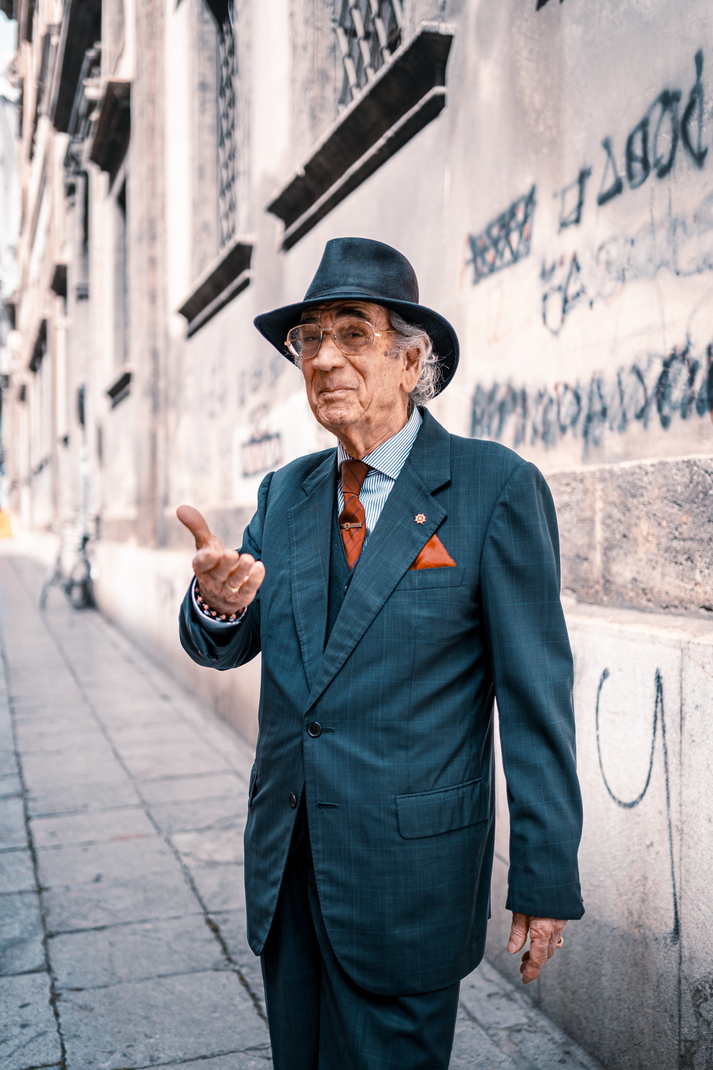 Elderly man in a suit and hat, standing on a graffiti-covered city street, gesturing with one hand.