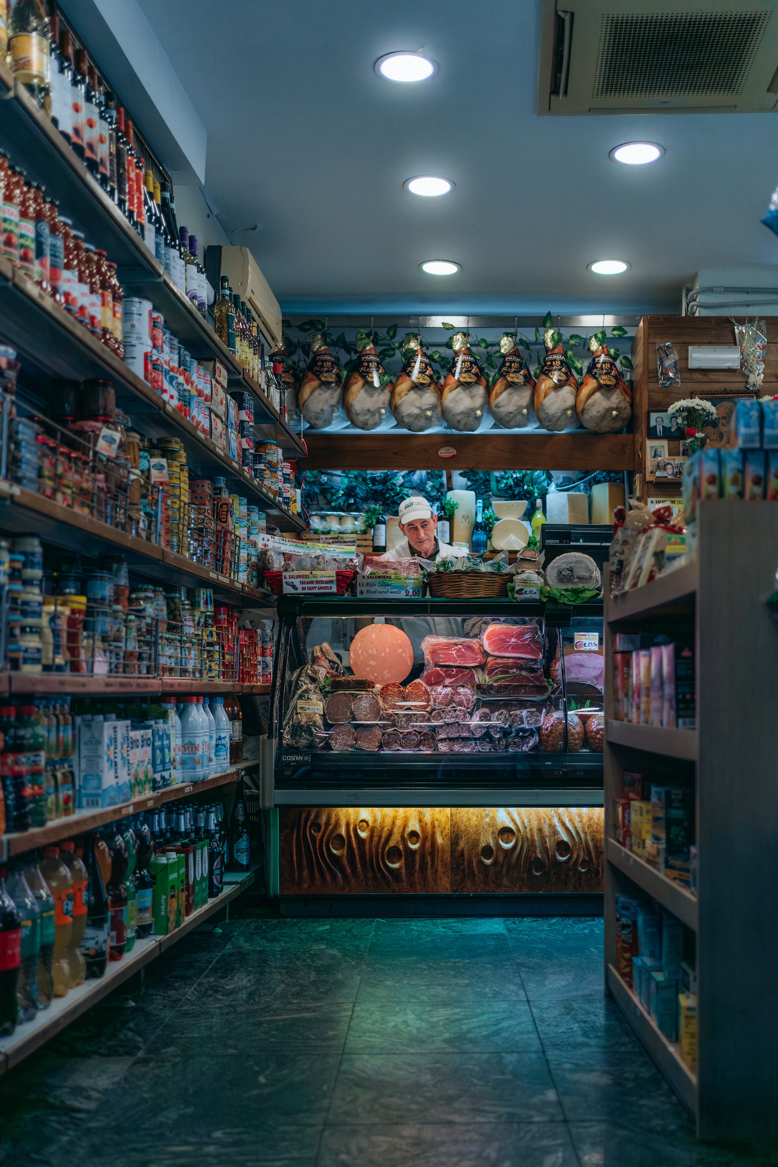 Interior of a small deli with shelves of groceries and a meat counter. A person stands behind the counter with various meats on display. Hanging cured meats and products are visible.