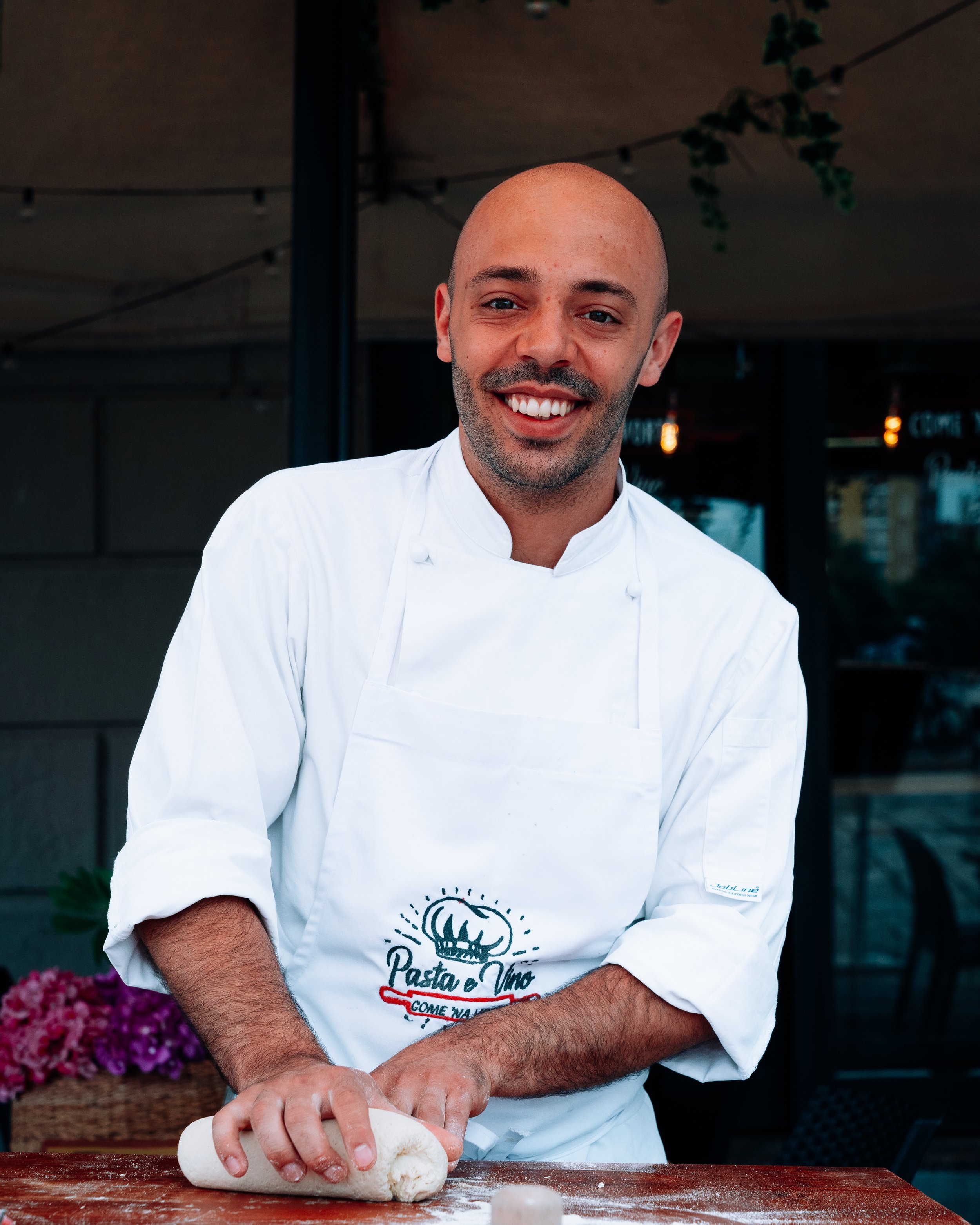 Smiling chef in a white apron kneading dough on a wooden table outdoors.