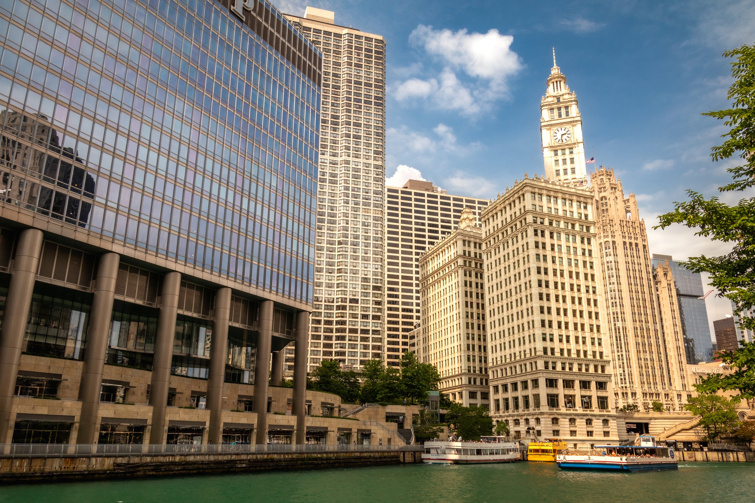 Chicago River view with skyscrapers, including historic Wrigley Building, and a tour boat, under a blue sky with clouds.