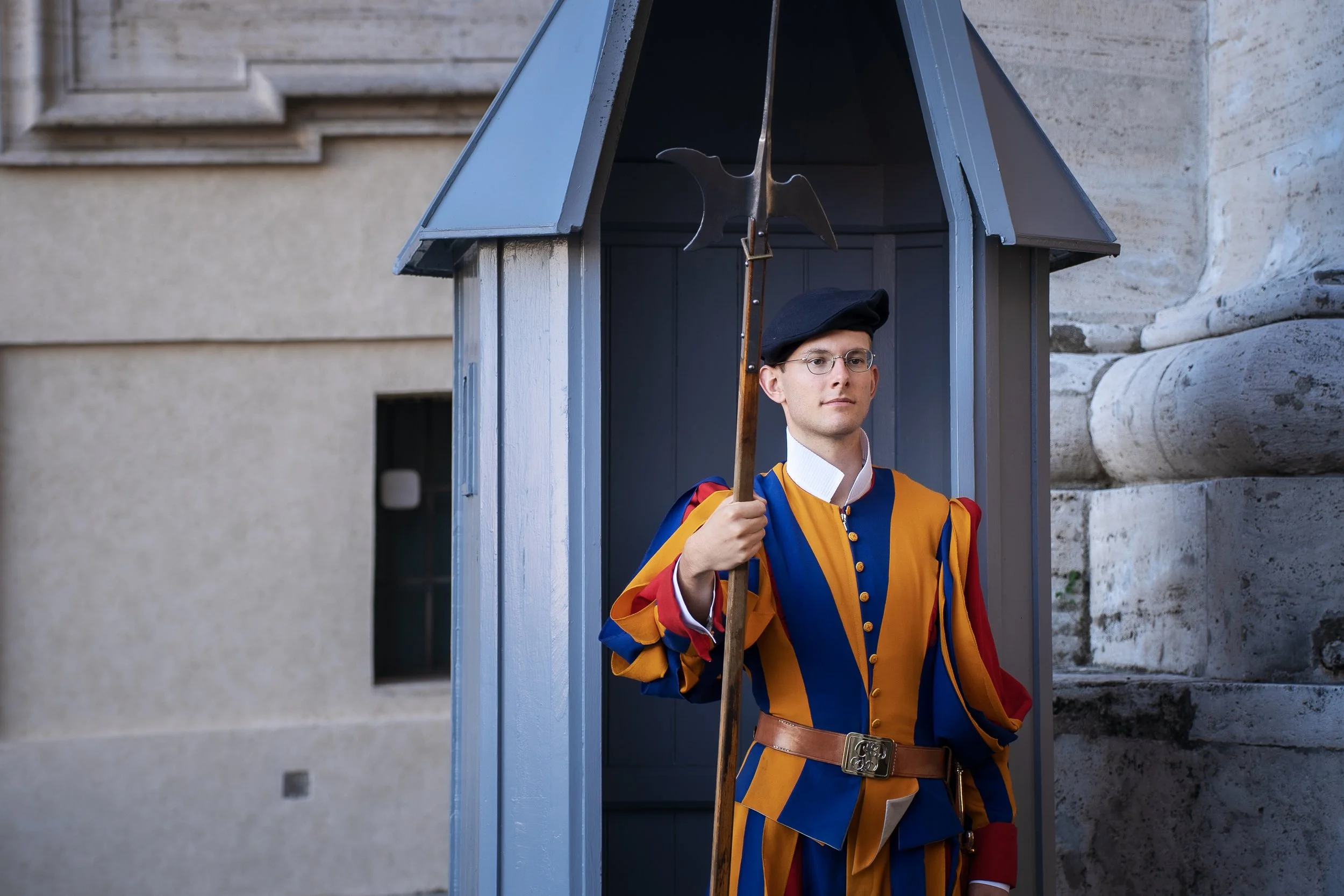 A ceremonial guard dressed in colorful, historical uniform standing inside a small guardhouse with a spear, in front of a stone building.