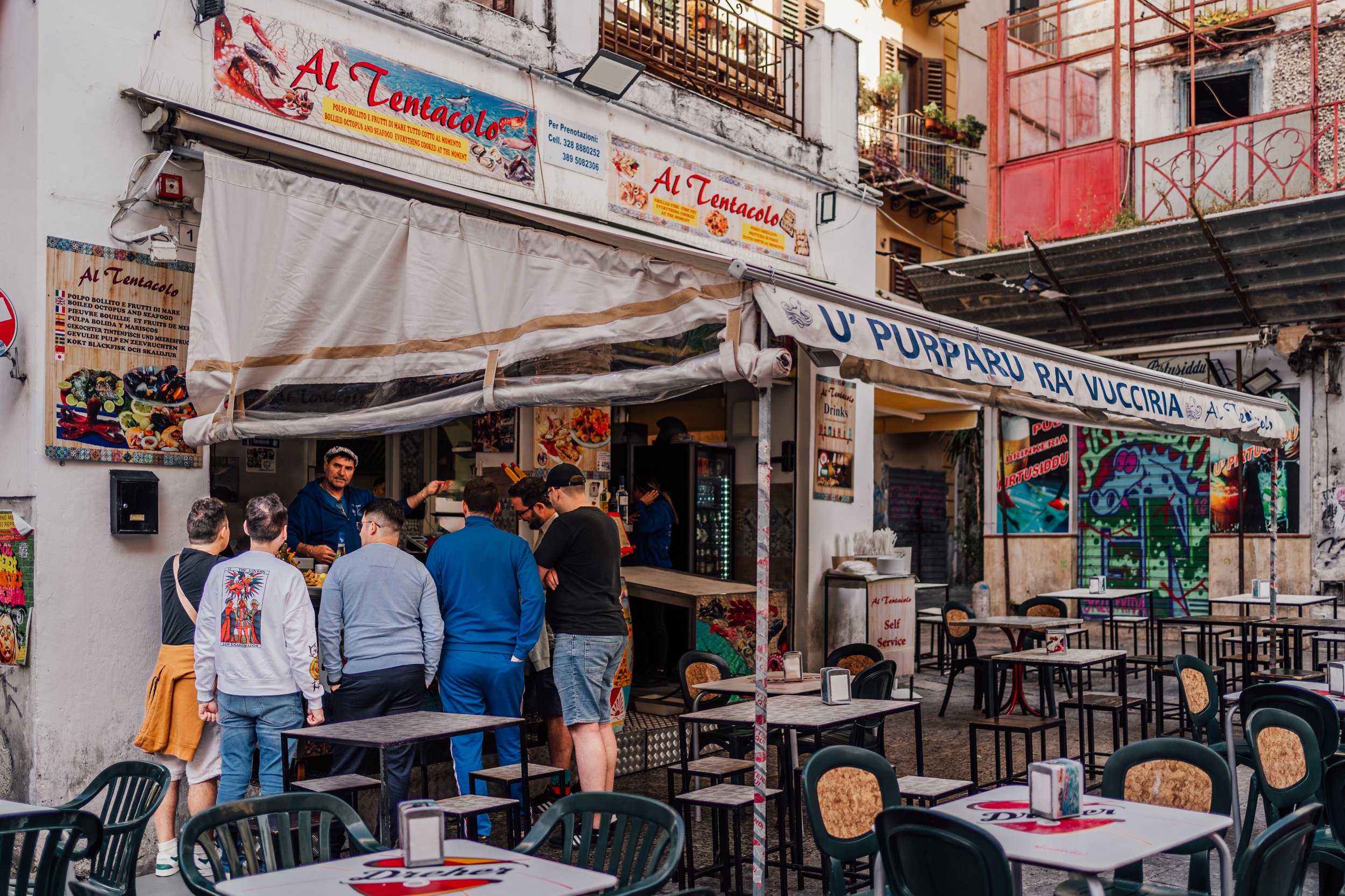 Outdoor restaurant in a narrow street, people ordering at a counter under a canopy labeled 'Al Tentacolo.' Tables and chairs are set up in front of the restaurant.
