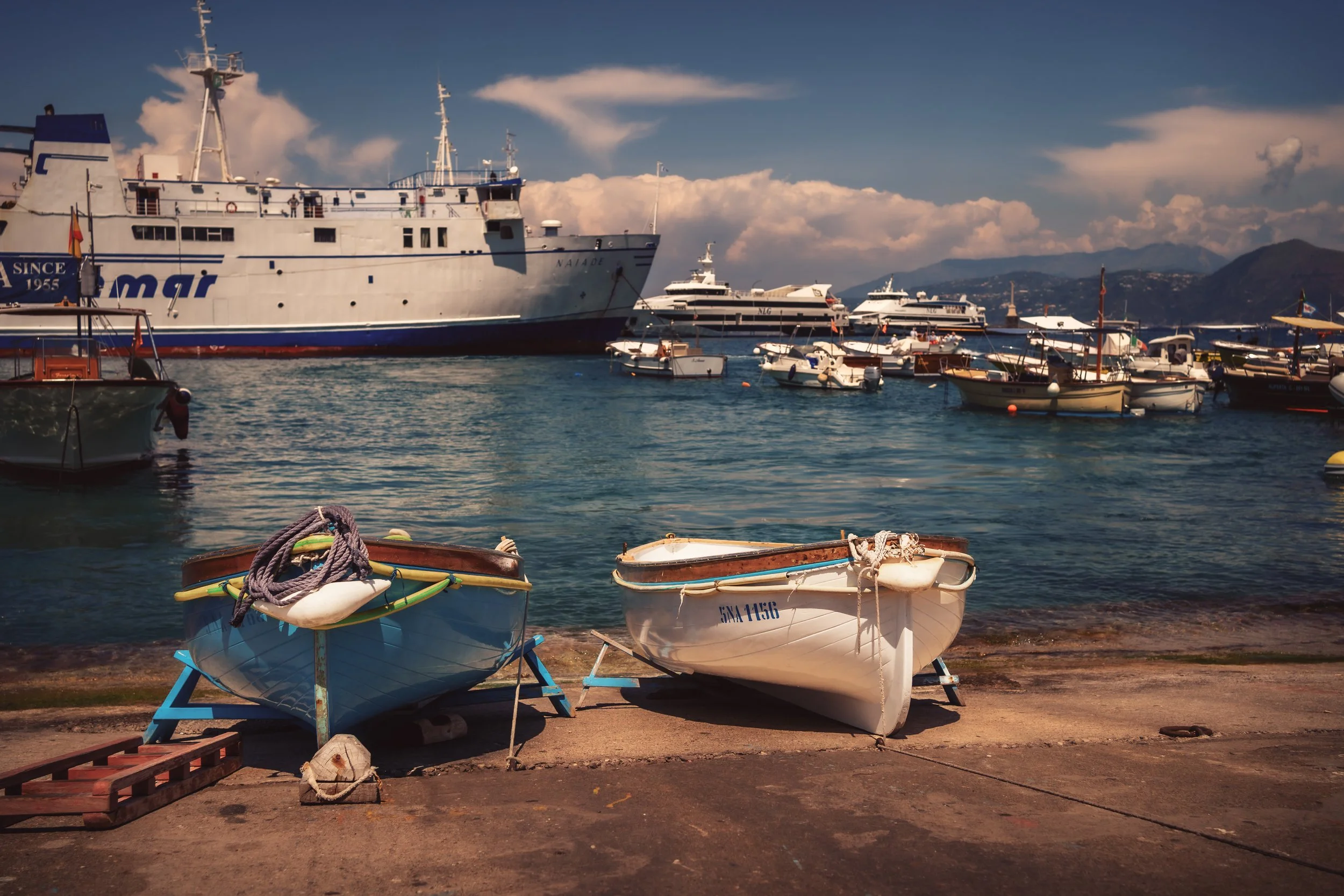 Two small boats on a concrete dock in front of a harbor filled with larger boats and yachts, with mountains in the background under a partly cloudy sky.
