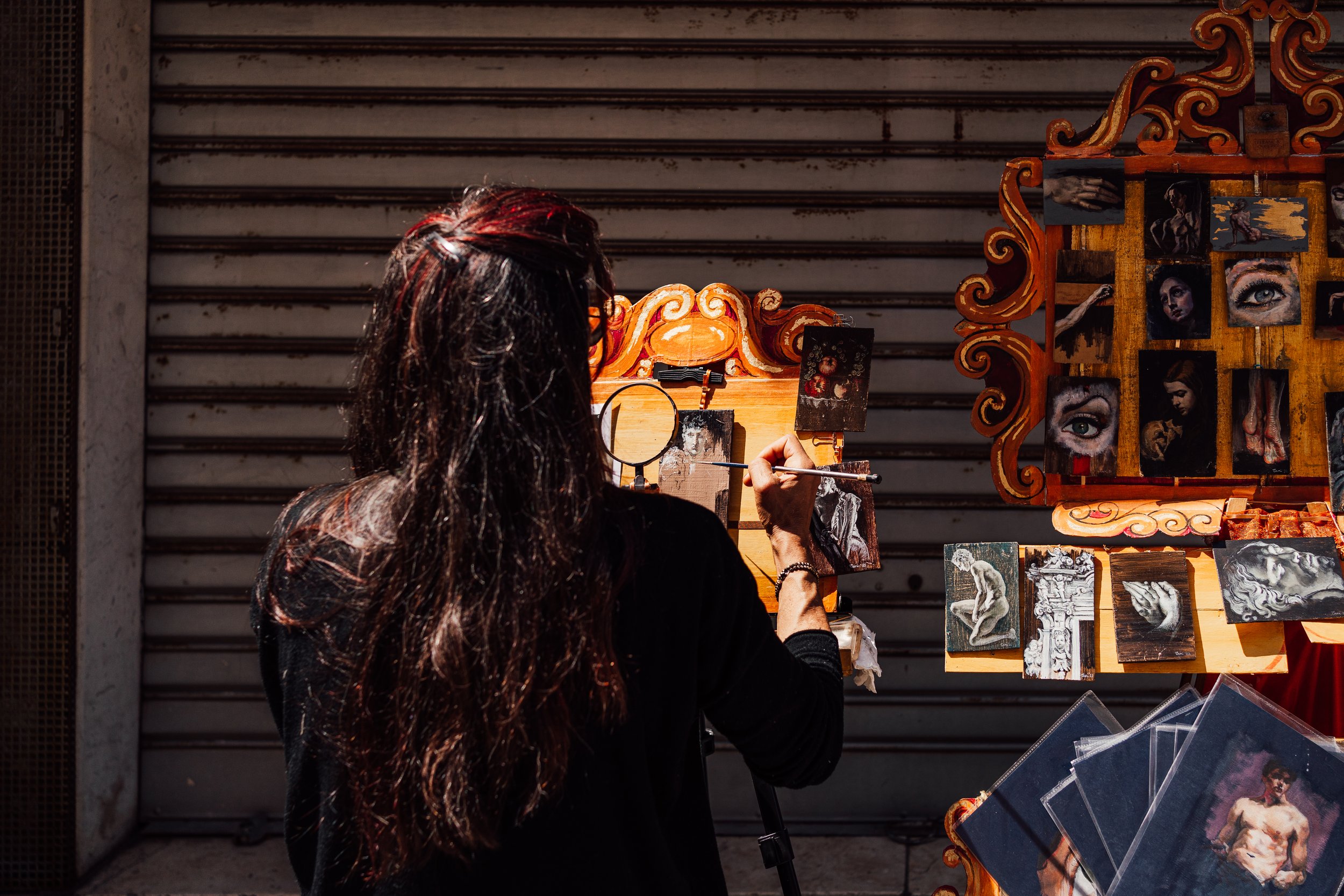 Artist painting at a street easel with small detailed artworks displayed on a colorful board.