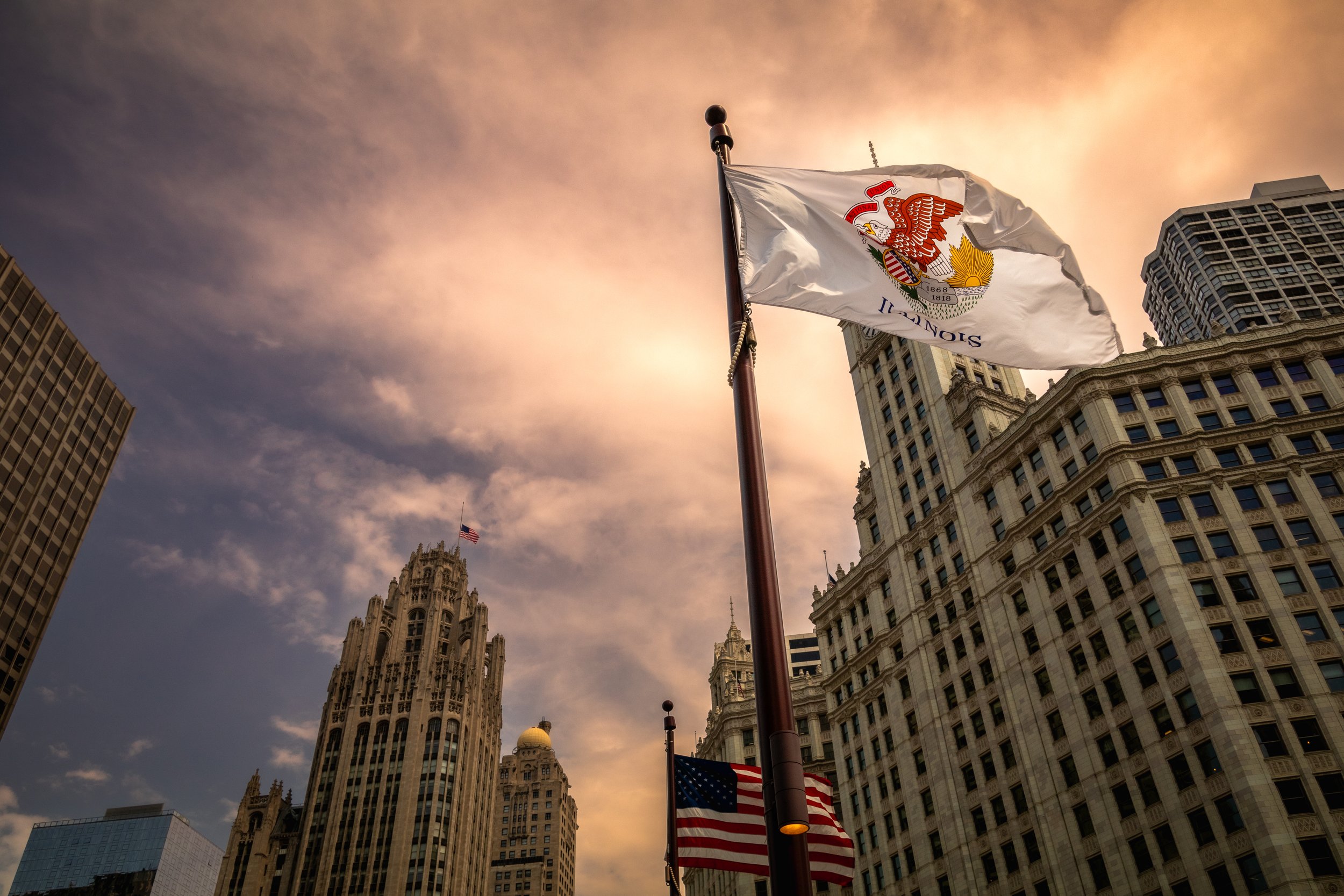 Illinois flag and American flag in front of Chicago skyscrapers with a dramatic sunset sky.