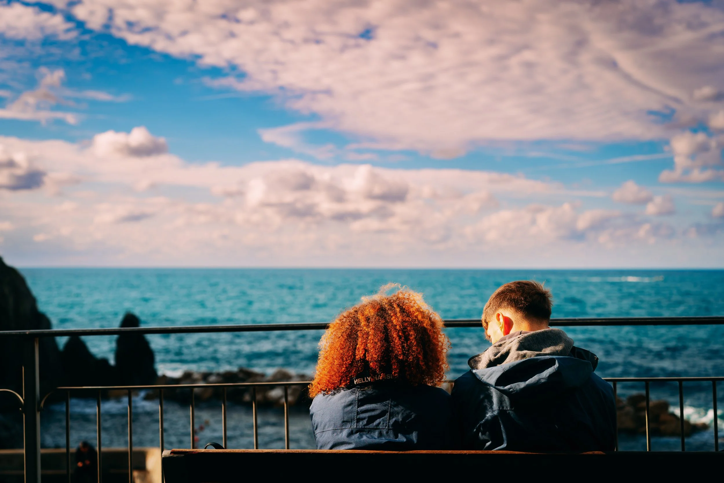 Two people sitting on a bench facing the ocean, with a sunset sky and clouds above, a rocky shoreline, and safety railing in the foreground.