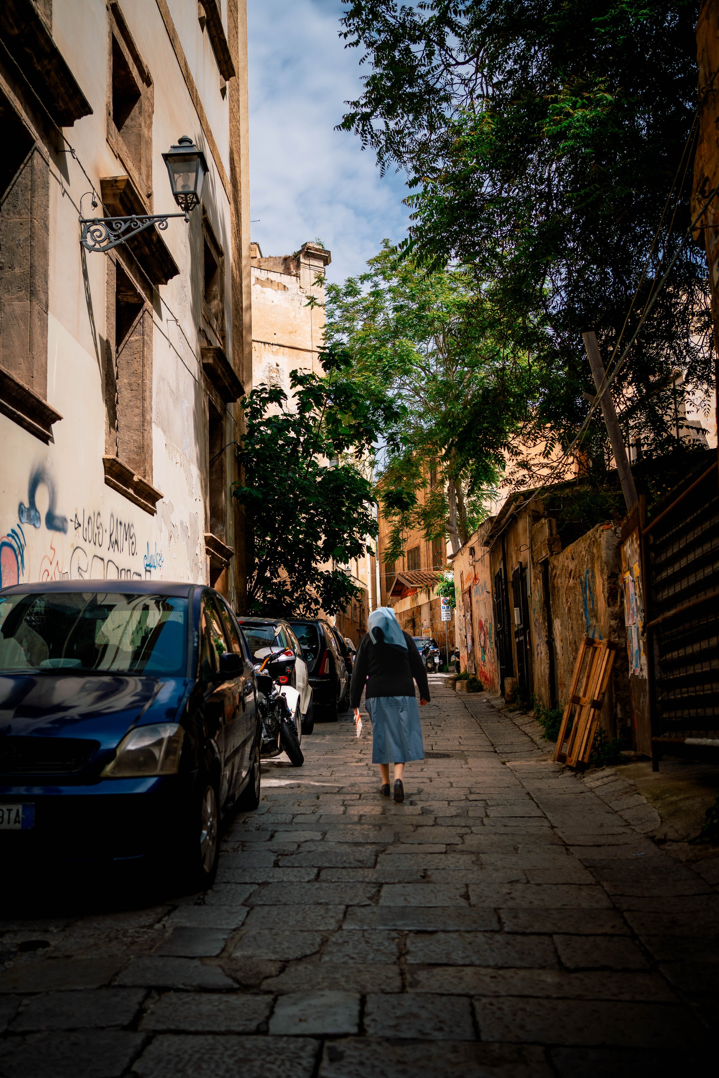 A narrow cobblestone alley with parked cars and a person walking, wearing a long skirt and blue headscarf. The walls are covered with graffiti, and there is a street lamp attached to the building. Trees partially shade the area, and a clear blue sky