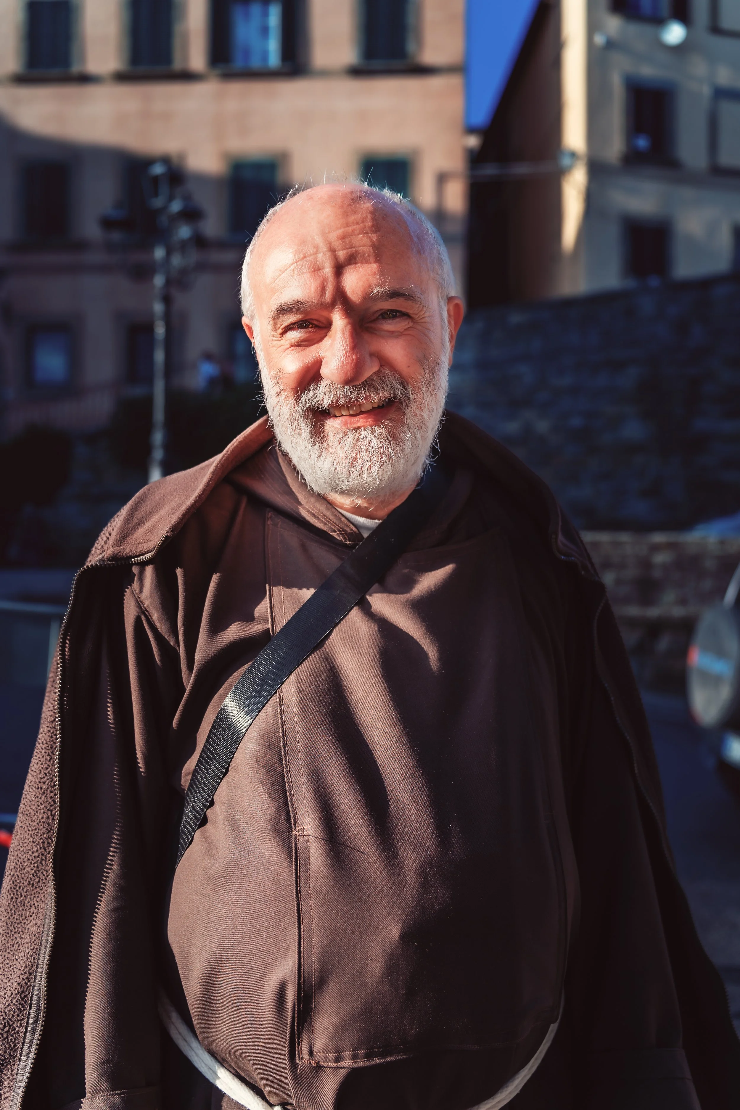A smiling elderly man with a white beard and bald head standing outdoors in urban sunlight, wearing a brown jacket and carrying a black strap across his chest.