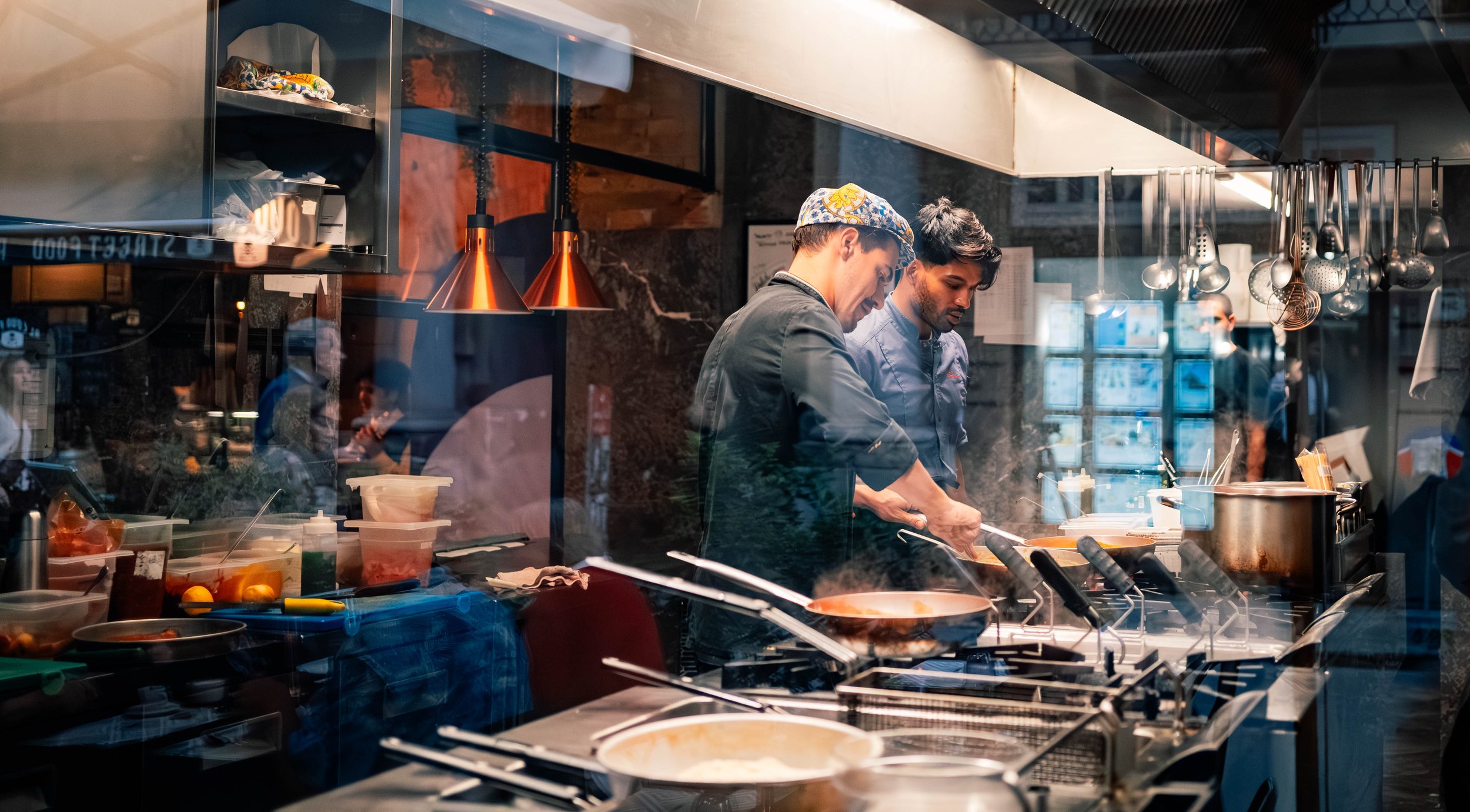 Two chefs cooking in a professional kitchen, with various utensils and ingredients on the countertops.