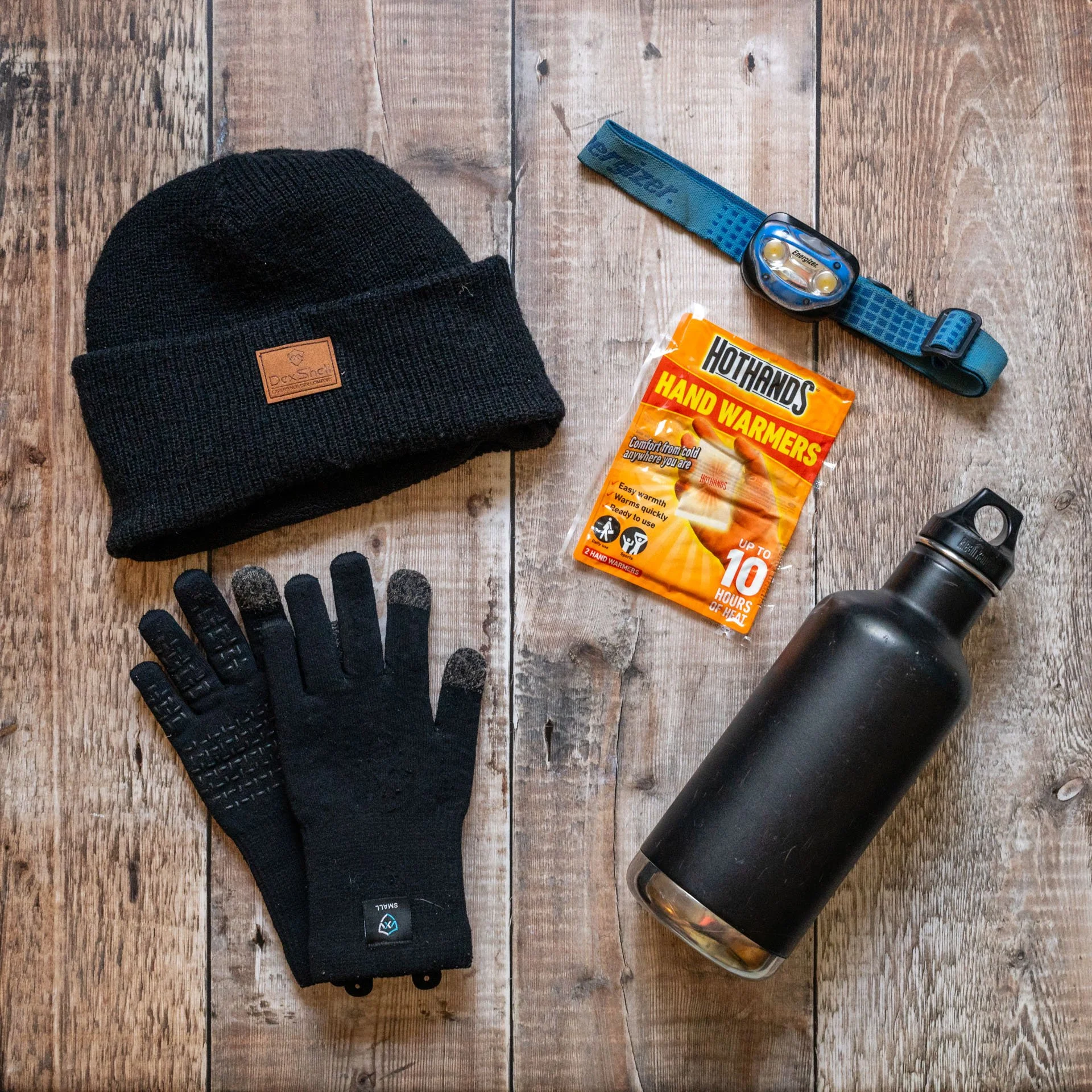 black hat and gloves, flask, head torch and handwarmers on a wooden floor