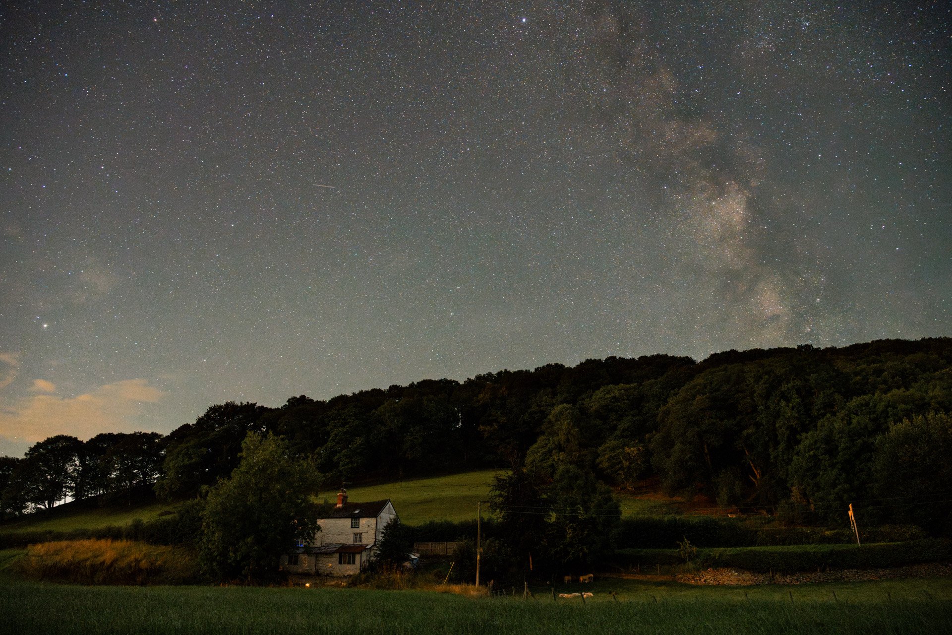 old, white cottage backing onto a feild at night with the milkyway and shooting stars in the sky