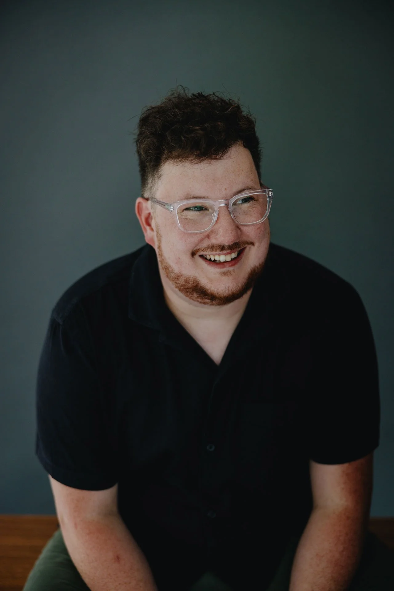 A smiling man with curly hair, glasses, and a beard, wearing a black shirt, sitting against a gray background.