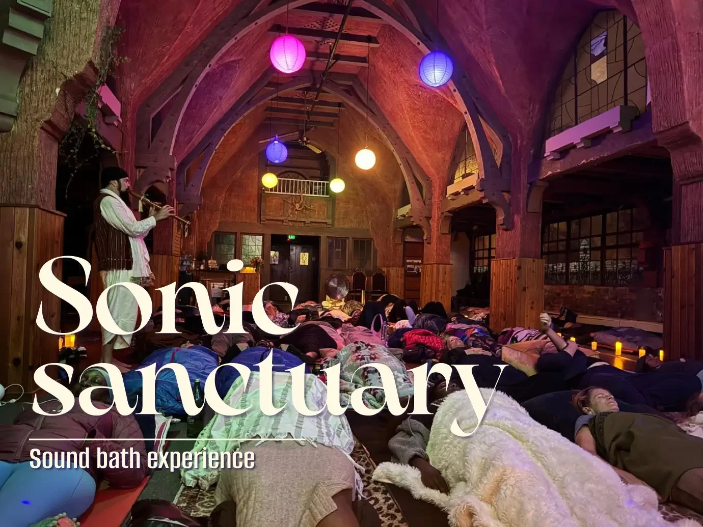 People lying on the floor with blankets in a wooden, vaulted-ceiling room, surrounded by colorful hanging paper lanterns, during a sound bath experience with a musician playing a flute.