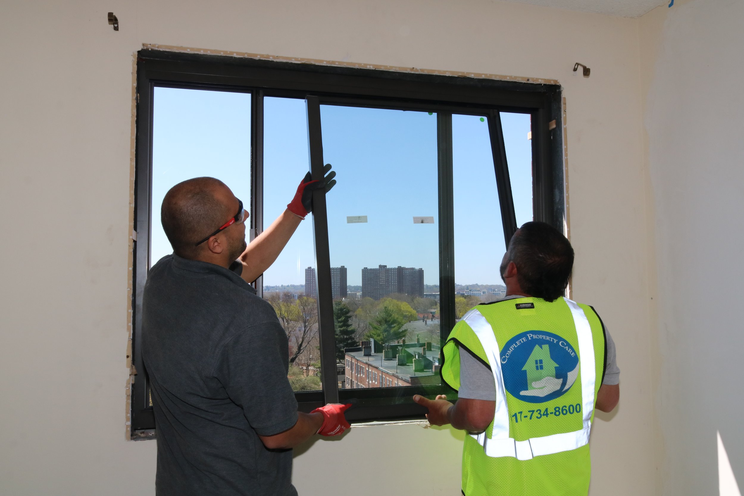Two construction workers installing a window in an unfinished room, with city buildings visible outside.
