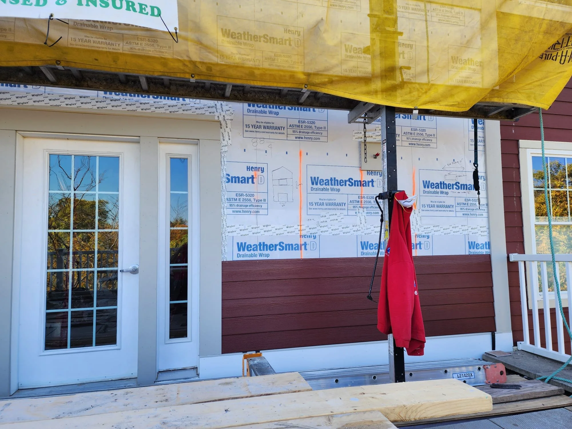 Construction site of a house showing a white door with multiple glass panes, red wooden wall, and weatherproofing material labeled 'WeatherSmart Drainable Wrap' on upper sections. A red cloth hangs on a standing pole, with a wooden platform nearby.