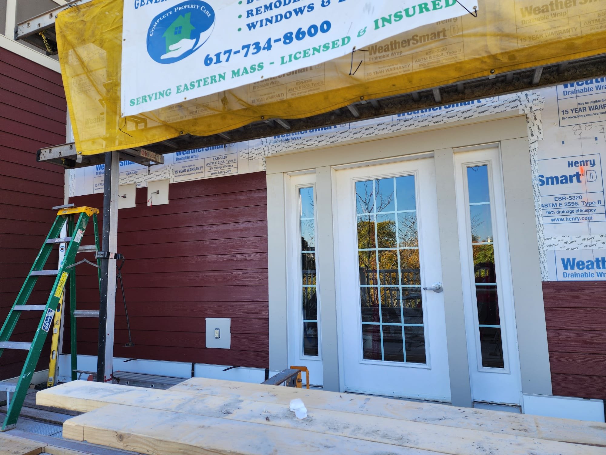 Construction site of a house with a red exterior, white door with glass panes, and a window. A yellow banner for a property care company hangs above, with a ladder and wooden planks in the foreground.