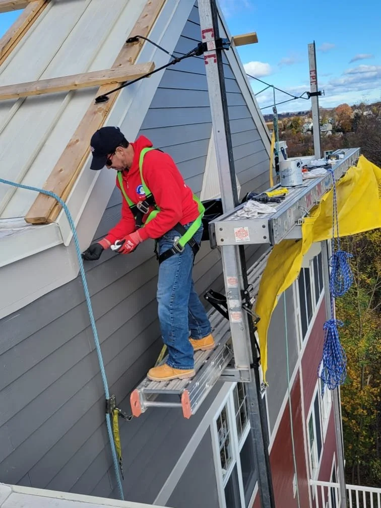 A construction worker in a red jacket, jeans, tan shoes, and a black cap is working on the side of a house, standing on a temporary scaffold. The house has blue-grey siding and is under construction or renovation, with exposed wooden beams and safety