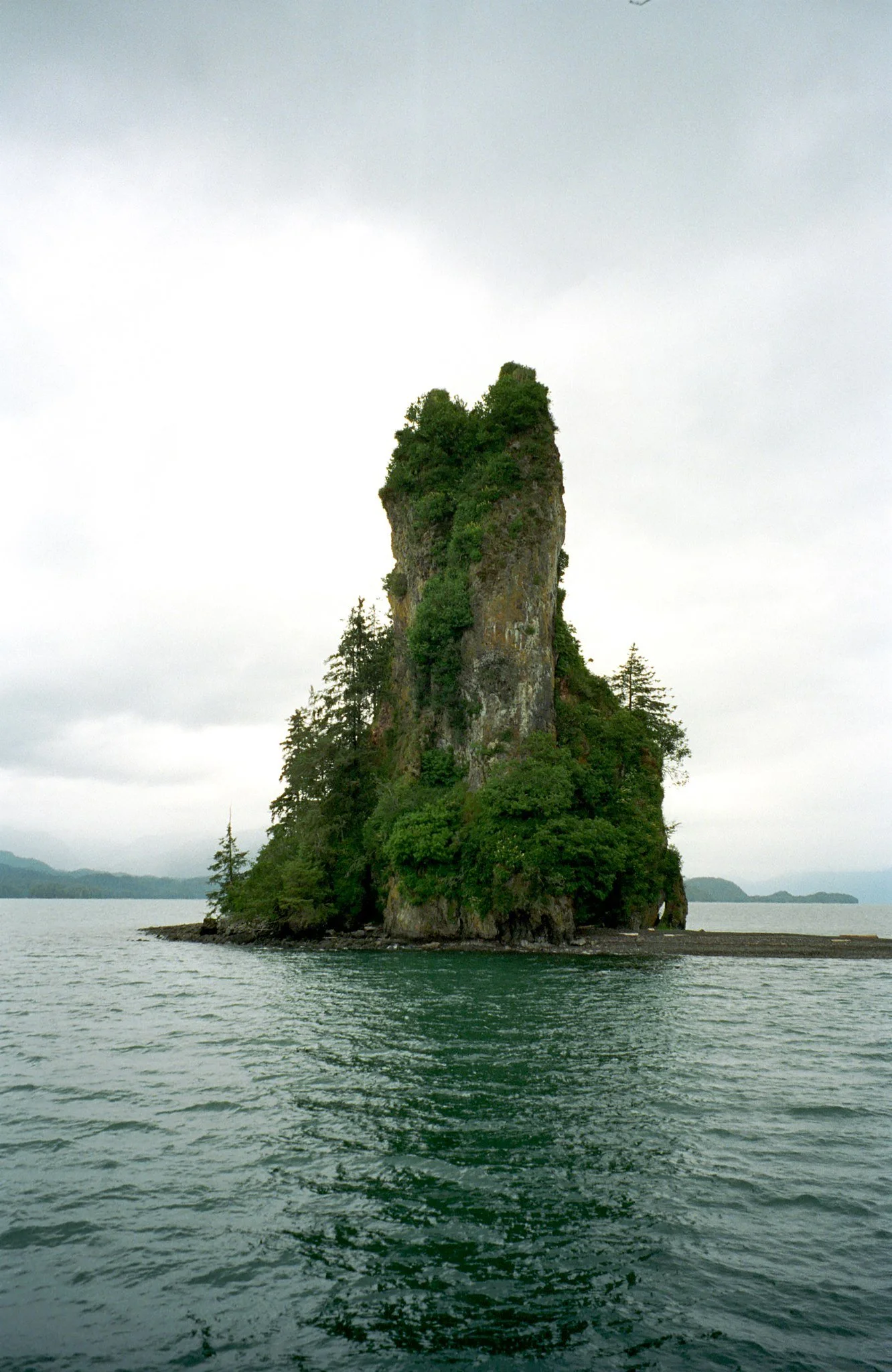 A tall, steeple-shaped rock formation covered with lush green trees rising out of the ocean, with distant landforms and an overcast sky in the background.