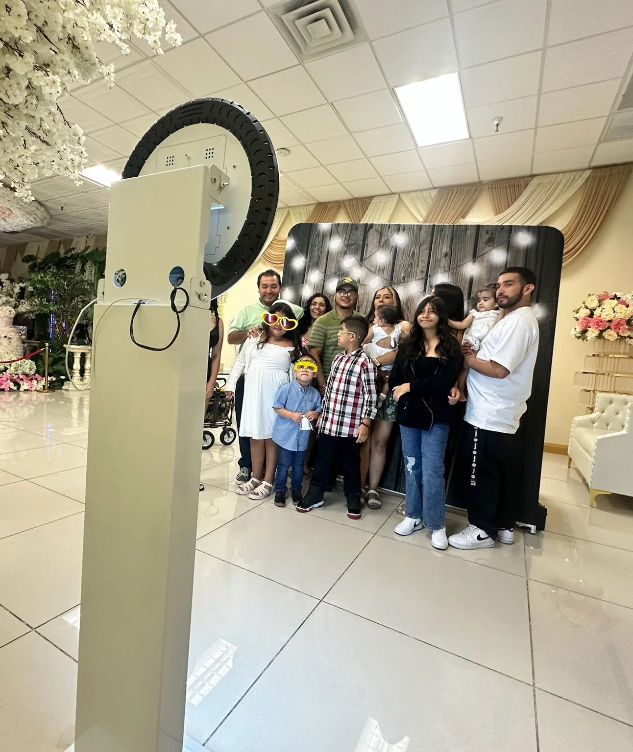 A family photo taken with a photo booth camera at an indoor event, with a group of people, including children, posing in front of a wooden background with string lights.