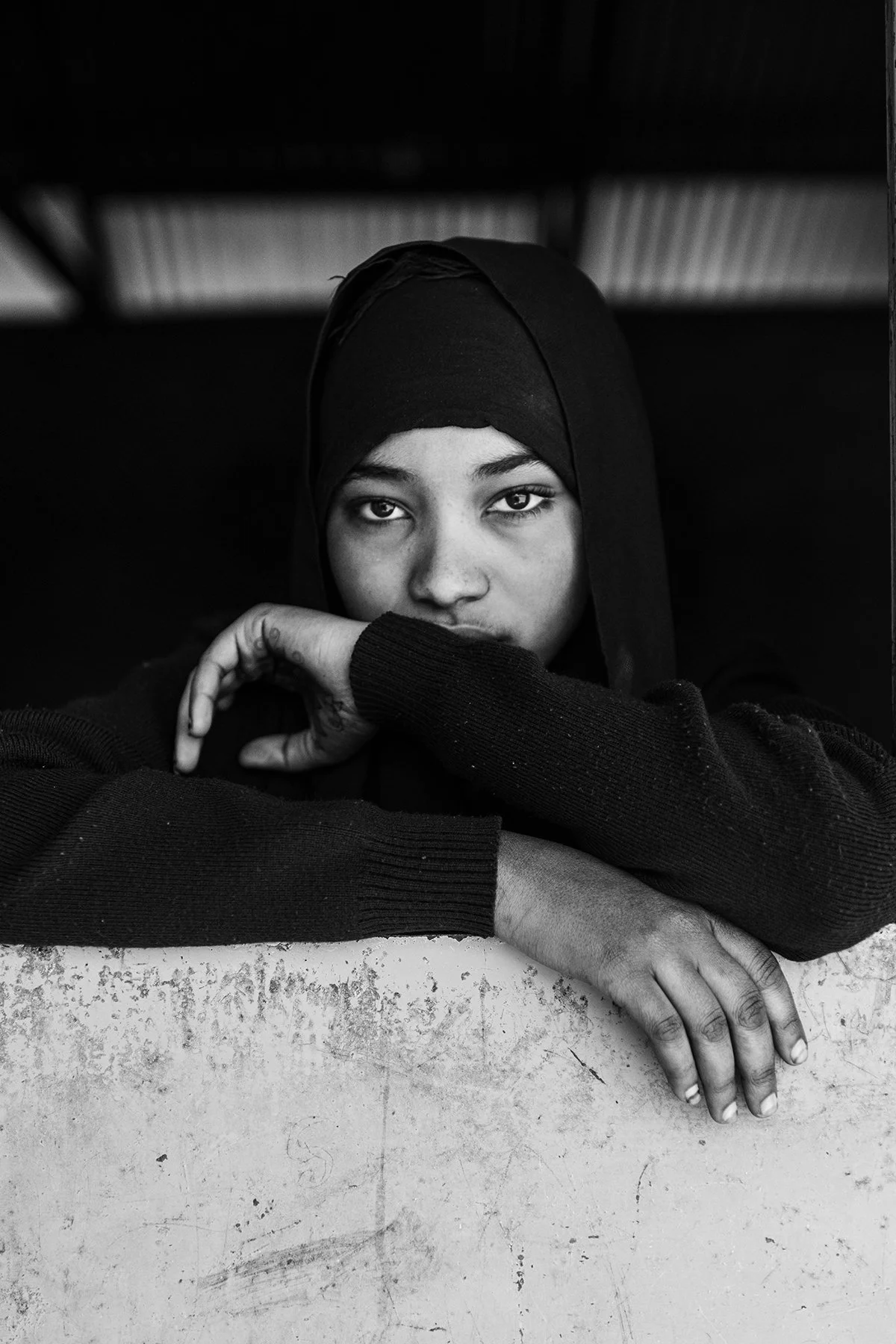 A young woman wearing a headscarf, resting her chin on her crossed arms, looking directly at the camera, in black and white.